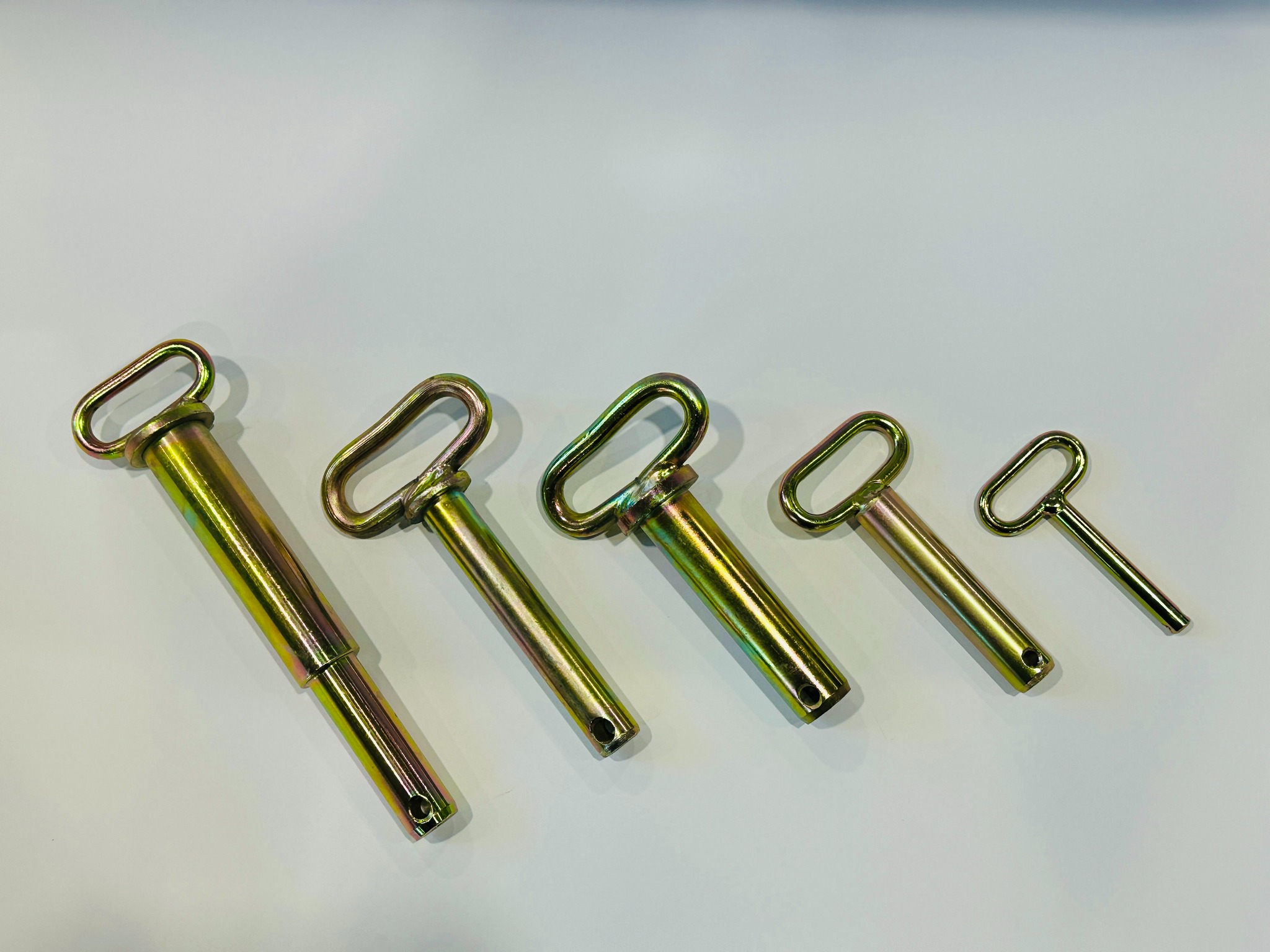 Szereg metalowych zawleczek o różnych rozmiarach, z charakterystycznym oczkiem, ułożonych na jasnym tle. Widoczne detale obróbki i metaliczny połysk.