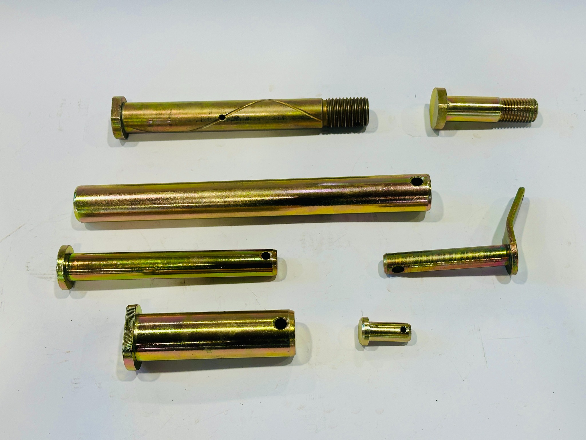 Zestaw metalowych, złoconych sworzni i trzpieni o różnych rozmiarach i kształtach, leżących na białym tle. Widoczne gwinty, otwory i charakterystyczne zakończenia.