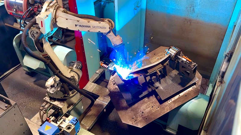 Zautomatyzowane stanowisko spawalnicze z robotem Yaskawa Motoman w akcji, iskry i niebieskie światło spawania, obróbka metalu w hali produkcyjnej.