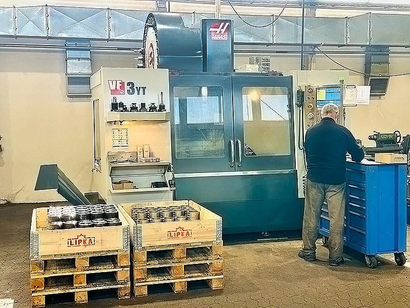 Praca przy maszynie CNC w warsztacie. Operator stoi przy niebieskim wózku narzędziowym. W tle widoczne dwie skrzynie z obrobionymi elementami i system wentylacji.