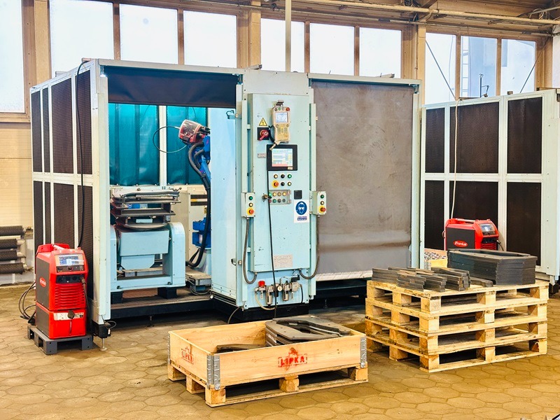 Duża, przemysłowa spawarka automatyczna z robotem spawalniczym, paletami i elementami metalowymi w hali produkcyjnej. Widoczne okna i oświetlenie przemysłowe.