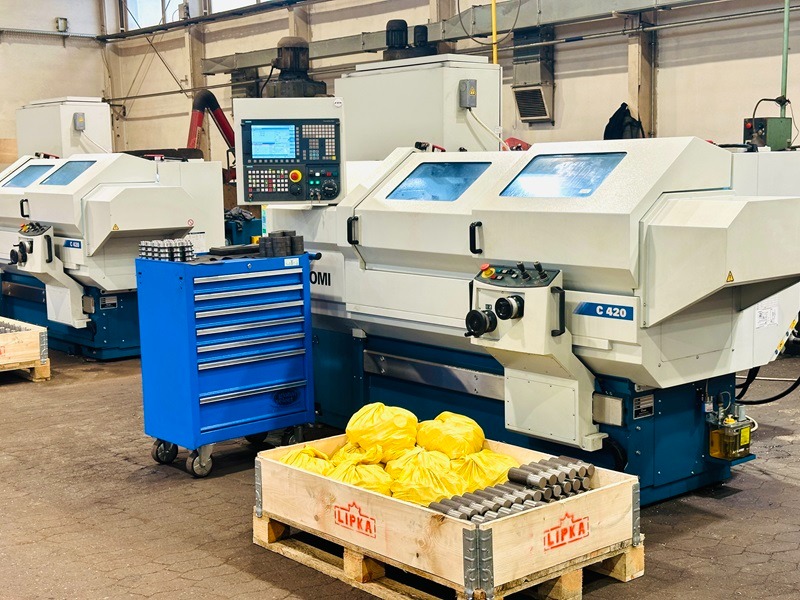 Dwa nowoczesne tokarki CNC w hali produkcyjnej, z niebieskim wózkiem narzędziowym i skrzynią z obrobionymi elementami na palecie. Widoczne detale maszyn i surowy charakter wnętrza.