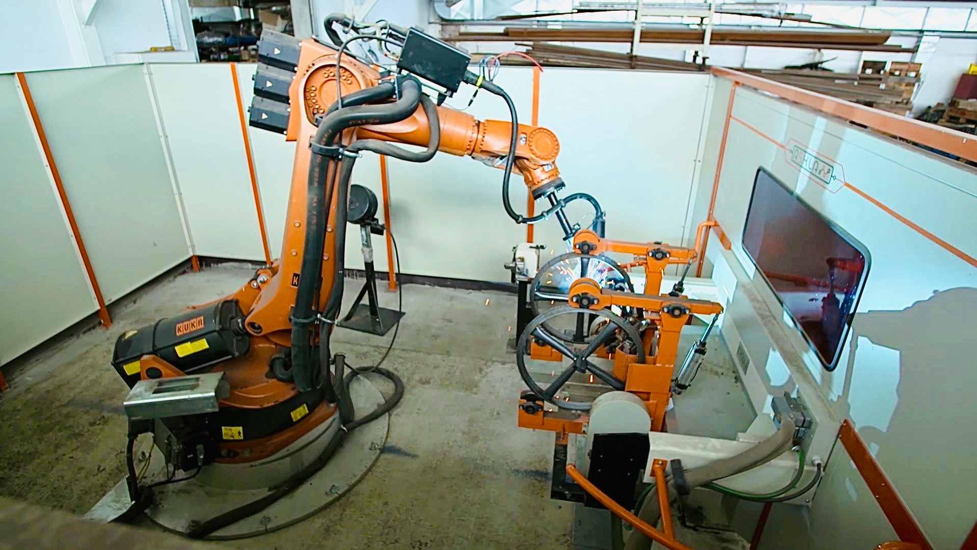 Pomarańczowy robot KUKA spawa metalowy element w osłonie. Widoczne iskry i wyposażenie ochronne. Ujęcie z góry, ukazujące precyzję i automatyzację procesu.