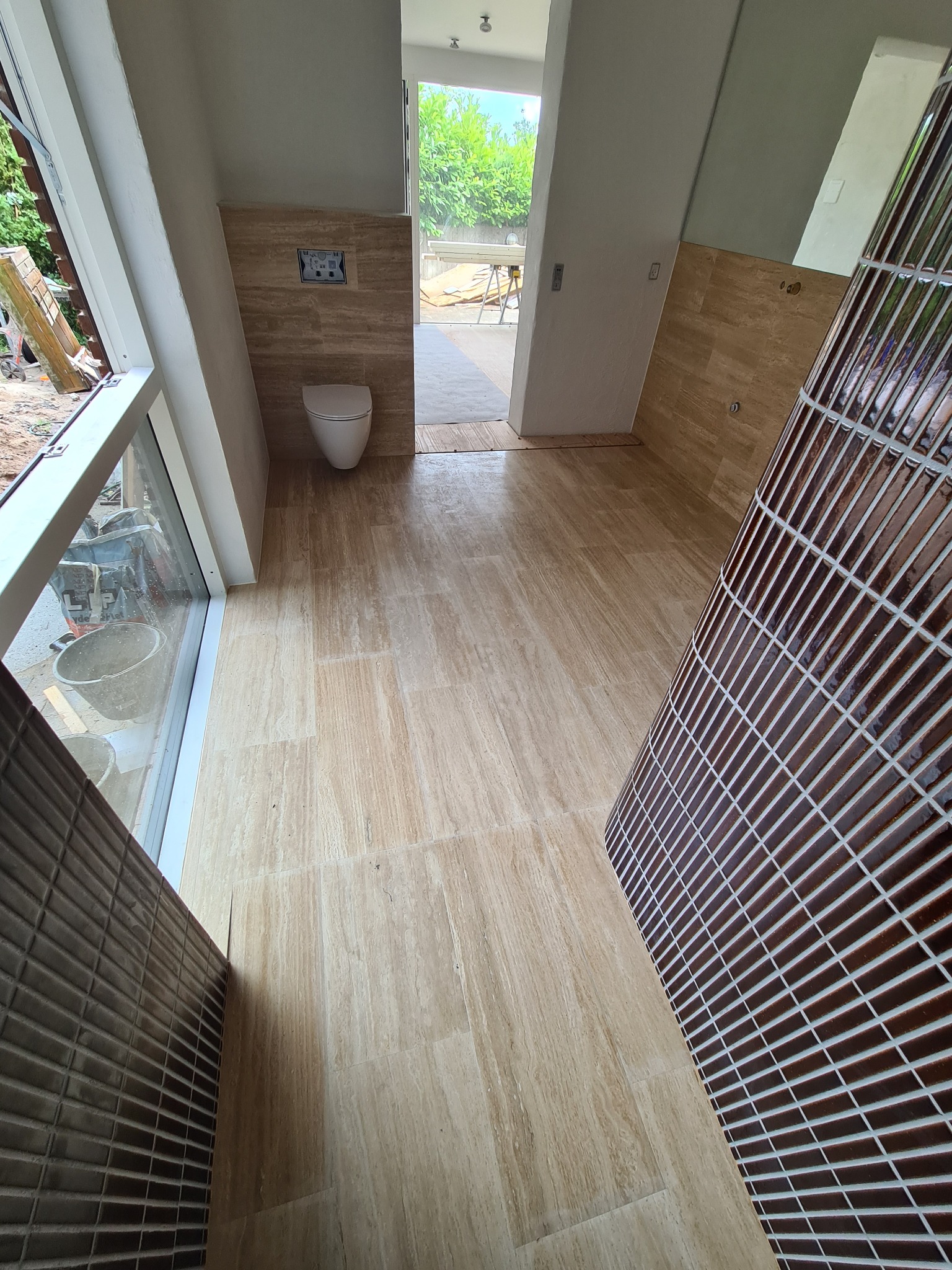 Wykończona łazienka z beżowymi płytkami imitującymi trawertyn na podłodze i ścianach. Uwagę przykuwa kolumna z brązowej mozaiki. Widoczna toaleta podtynkowa.