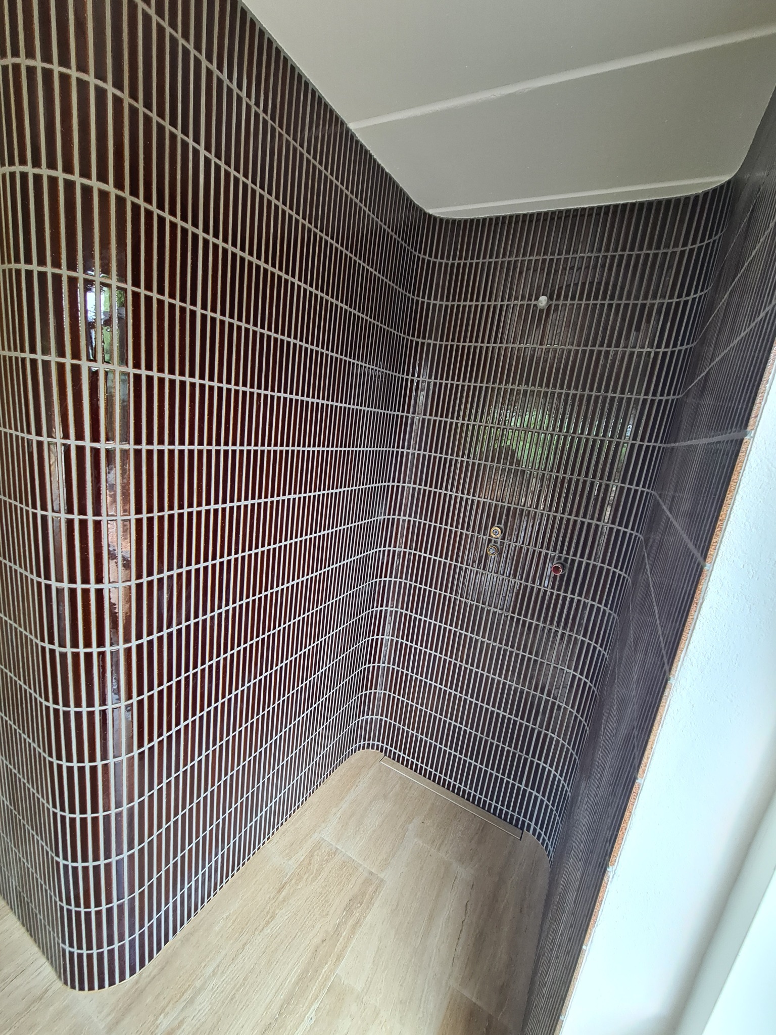 Nowoczesna kabina prysznicowa z brązowymi, pionowo ułożonymi płytkami typu 'cegiełka' i jasną podłogą imitującą drewno. Zaokrąglone narożniki ścian.