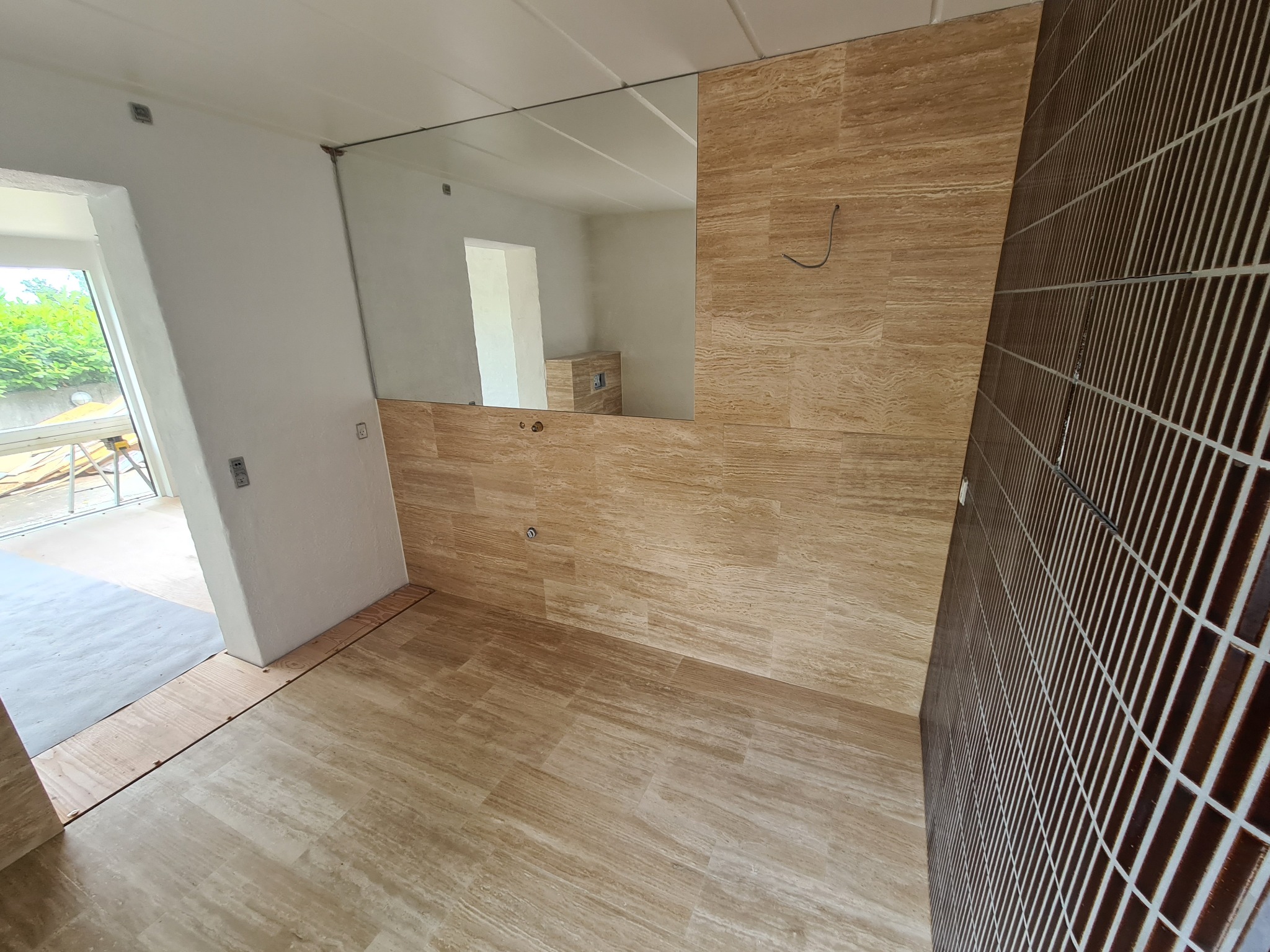 Wykończona łazienka z beżowymi płytkami imitującymi drewno na ścianach i podłodze, oraz brązowymi płytkami w kratkę na jednej ze ścian. Lustro bez ramy na ścianie.