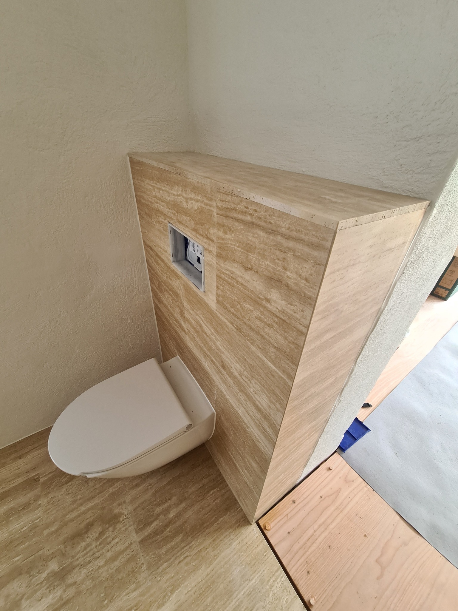 Wykończona toaleta z zabudową geberit z płytek imitujących trawertyn, widok z góry. Beżowe płytki na podłodze i ścianie. Otwór rewizyjny w zabudowie.