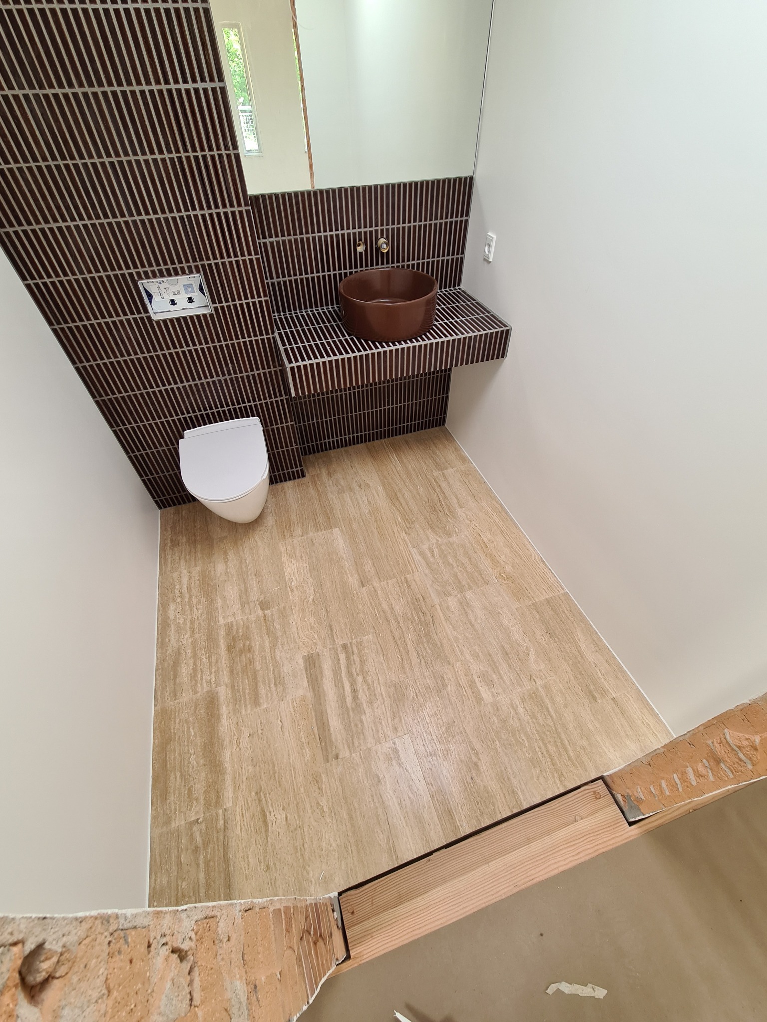 Nowoczesna łazienka po remoncie: brązowe płytki imitujące lamele na ścianie, umywalka w odcieniach brązu, beżowa podłoga z płytek. Widok z góry, fragment muru w rogu.