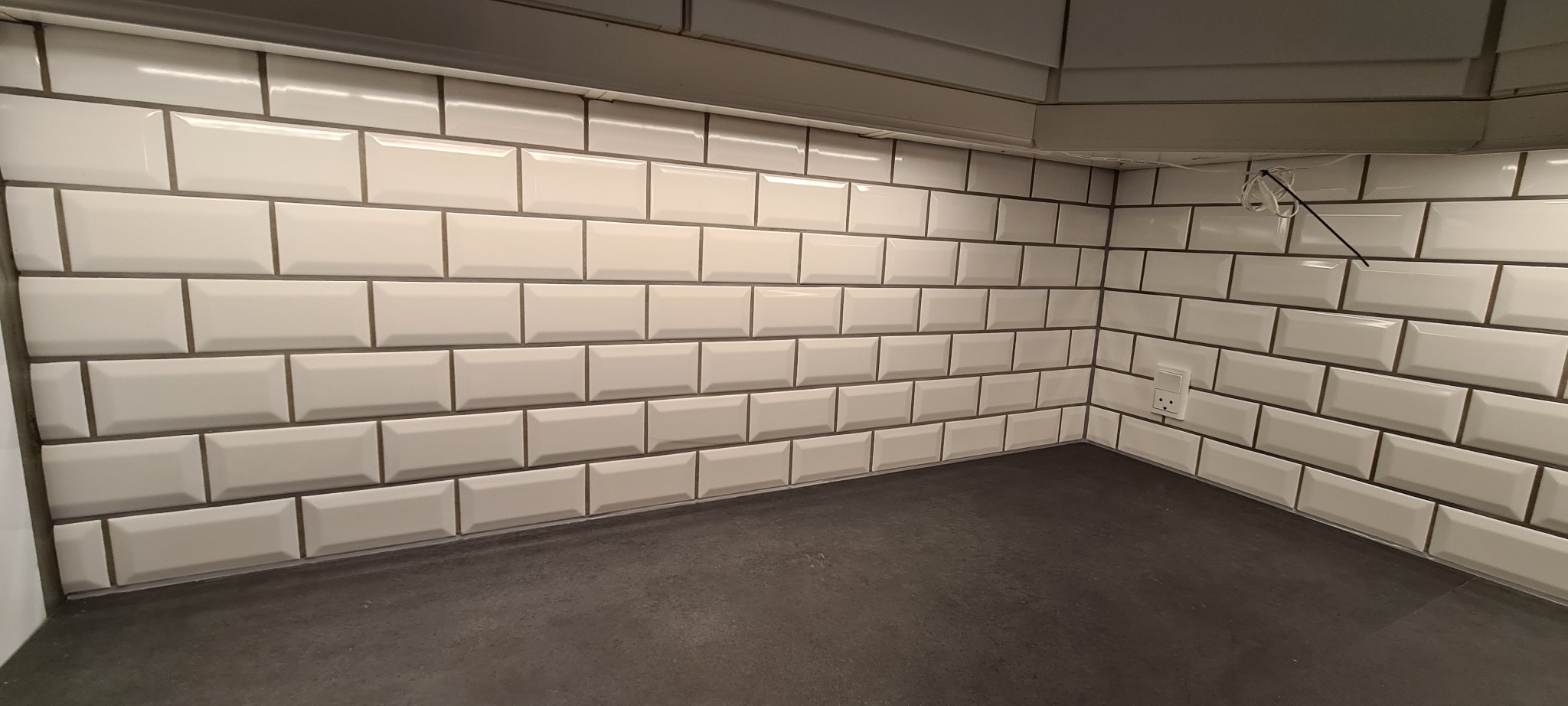 Białe, błyszczące płytki typu metro ułożone na ścianie nad blatem kuchennym. Widoczny fragment szafki wiszącej i gniazdko elektryczne. Minimalistyczny design.