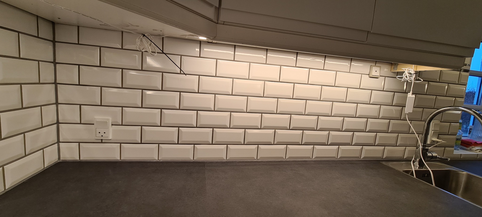 Białe, prostokątne płytki ułożone w cegiełkę na ścianie nad blatem kuchennym. Widoczne gniazdka elektryczne i przewody oświetleniowe pod szafkami.