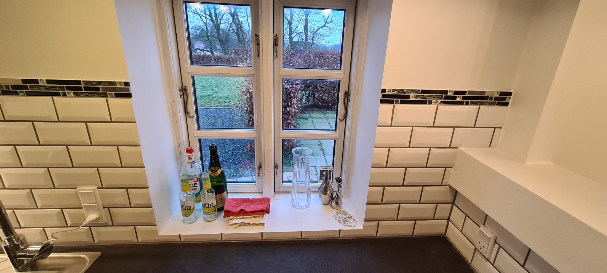 Wykończona kuchnia z białymi, błyszczącymi płytkami typu metro i dekoracyjnym pasem mozaiki. Na parapecie okna stoją butelki i dekoracje.