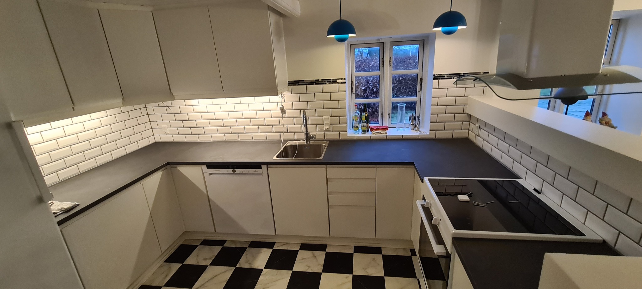 Nowoczesna kuchnia z białymi szafkami, czarnym blatem i białą glazurą typu metro. Uwagę przykuwają niebieskie lampy wiszące i podłoga w czarno-białą szachownicę.