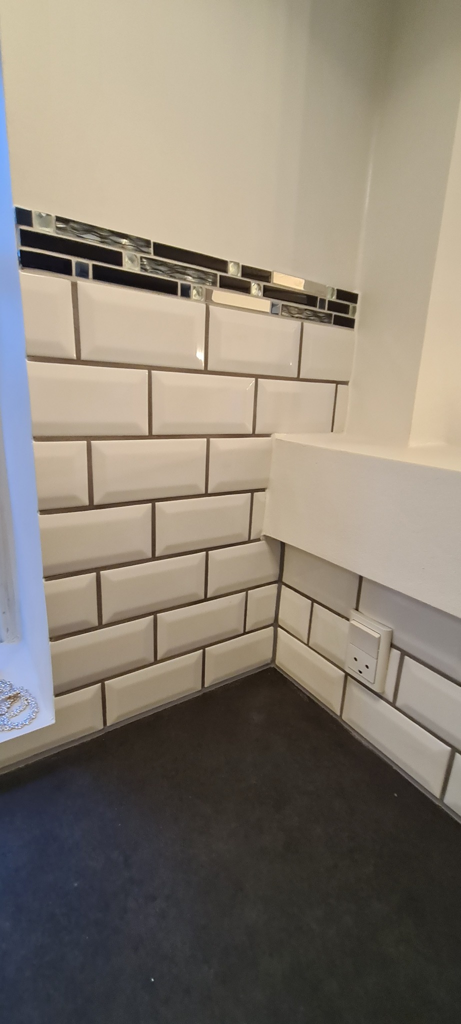 Wykończona ściana z białych, błyszczących płytek typu metro, z dekoracyjnym pasem mozaiki. Widoczny fragment blatu i gniazdko elektryczne. Ujęcie z dołu, blisko ściany.