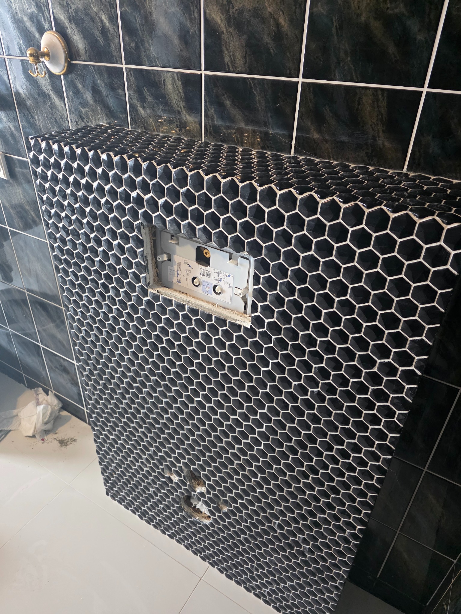 Obudowa stelaża WC wykończona czarną mozaiką heksagonalną z białymi fugami, widoczny fragment instalacji. Na ścianie czarne płytki imitujące marmur.