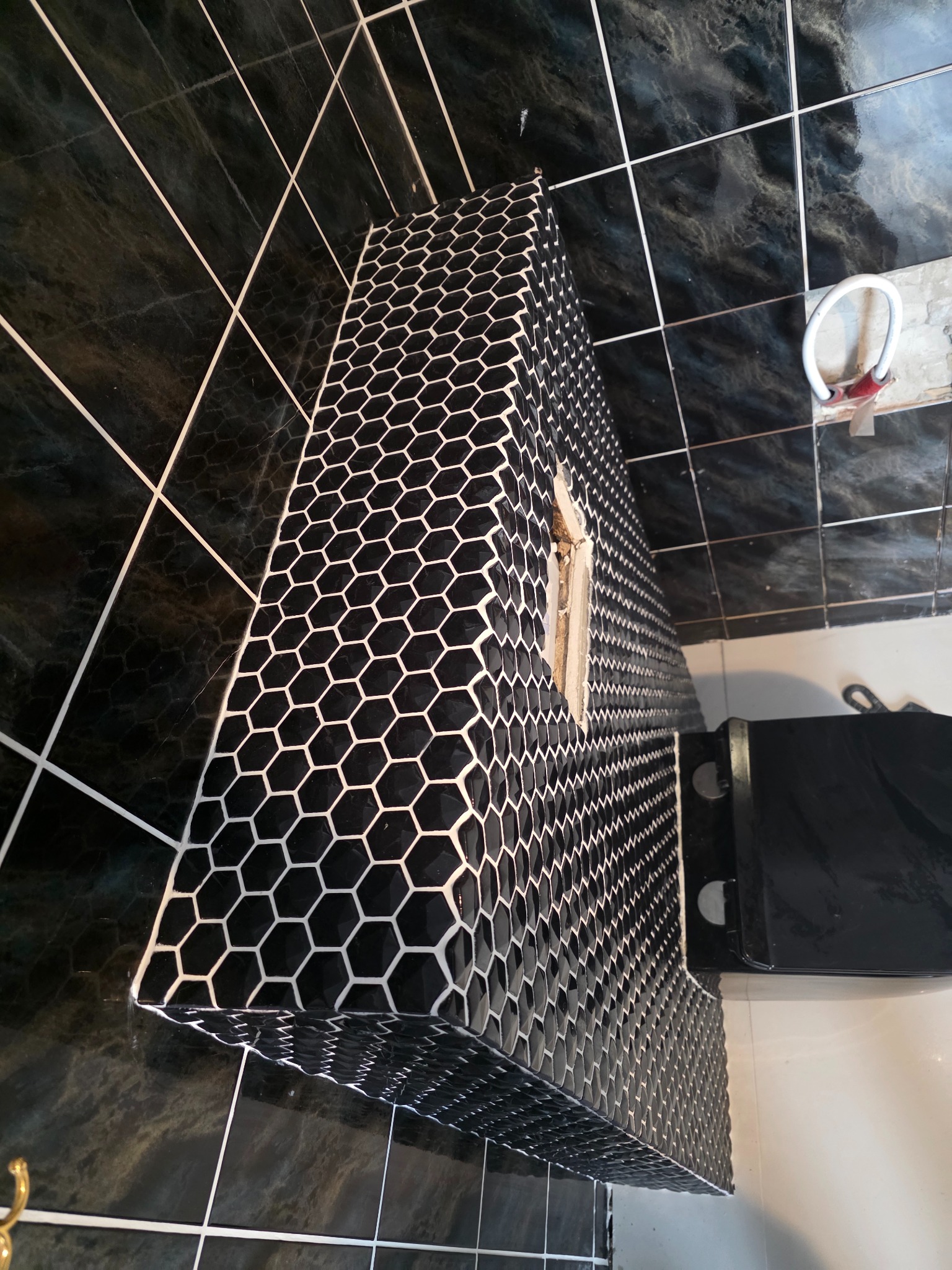 Wykończona obudowa umywalki z czarno-białej mozaiki heksagonalnej w łazience z ciemnymi, marmurkowymi płytkami. Widoczny fragment toalety i akcesoria łazienkowe.
