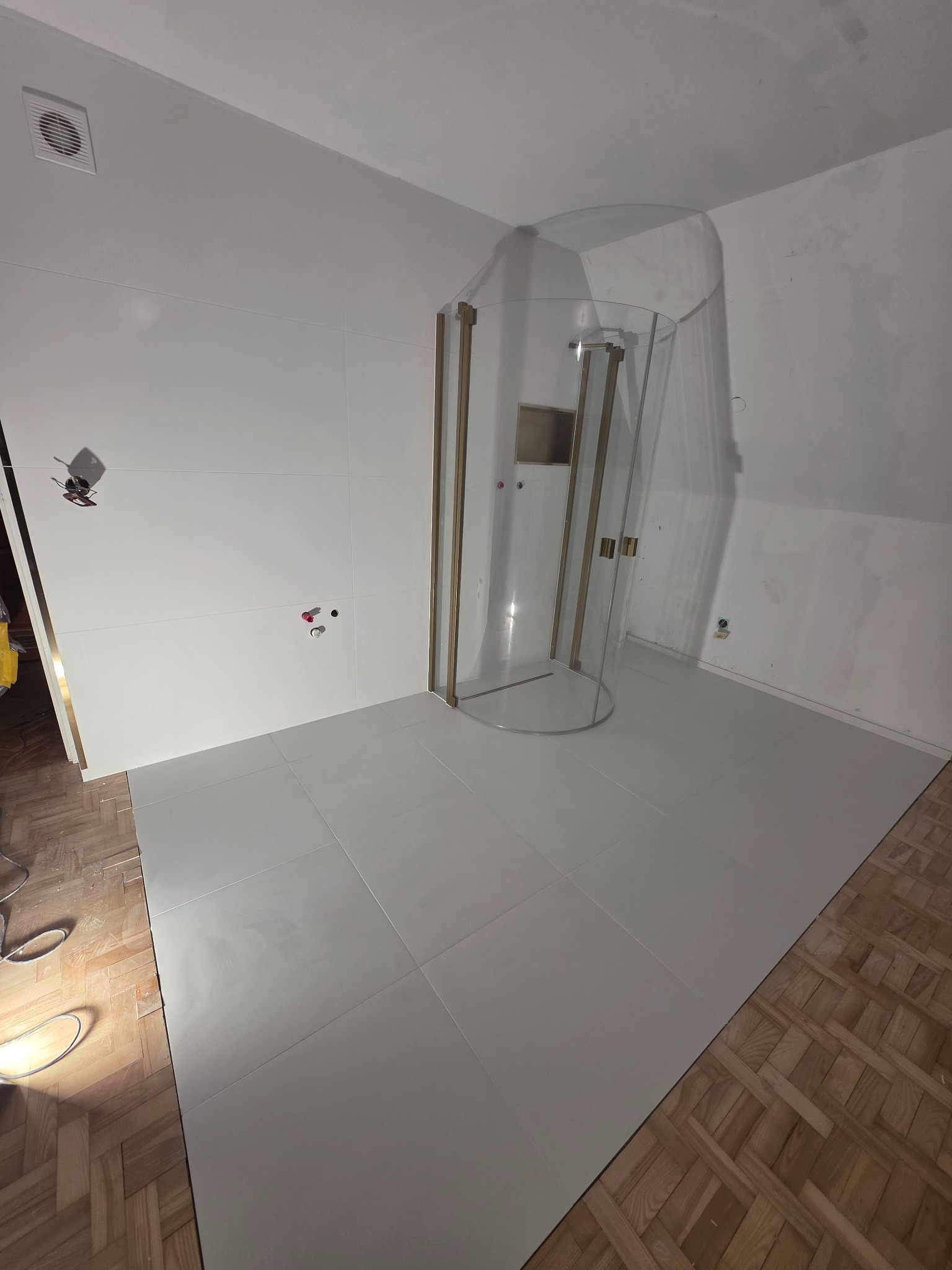 Nowoczesna kabina prysznicowa z złotymi detalami, zamontowana na jasnych płytkach podłogowych. Ściany wyłożone białymi, dużymi płytkami. Częściowo widoczna stara podłoga.