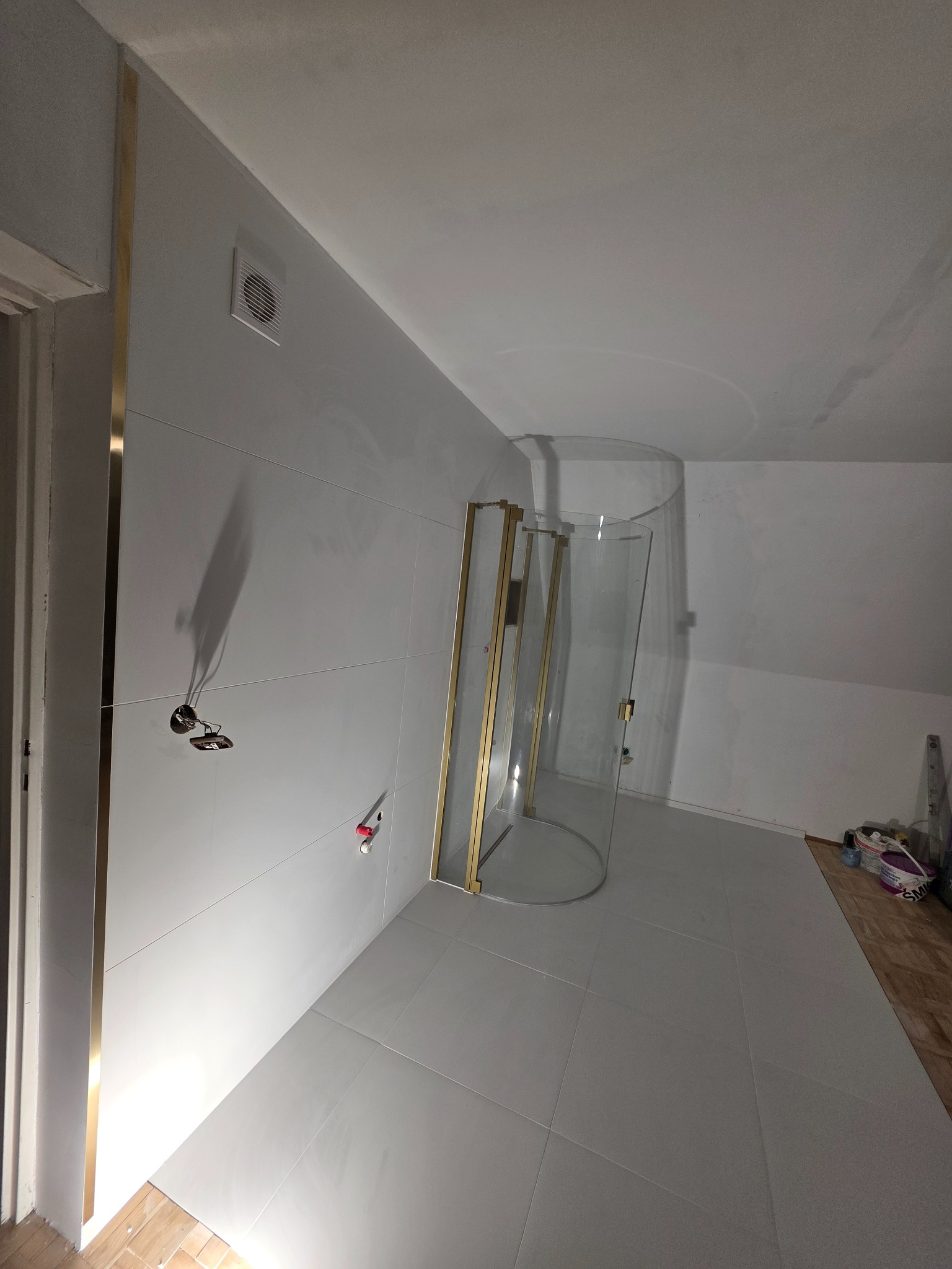 Wykończona łazienka na poddaszu z kabiną prysznicową typu walk-in z elementami w kolorze złotym, białe płytki na ścianach i podłodze, widoczne przygotowane instalacje.