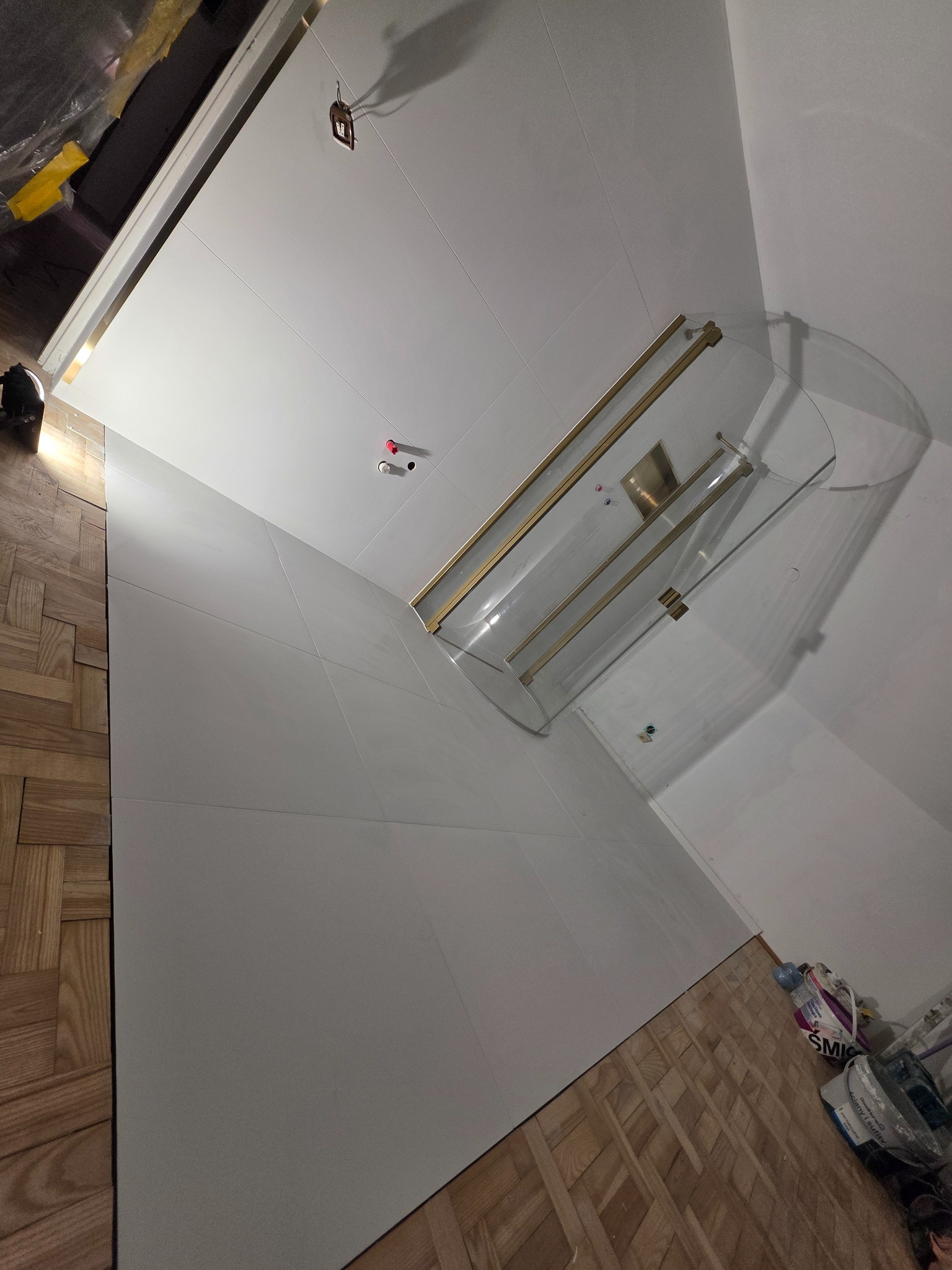 Białe płytki ścienne i podłogowe w łazience z kabiną prysznicową ze złotymi detalami. Parkiet ułożony w jodełkę. Widoczne elementy instalacji i akcesoria łazienkowe.