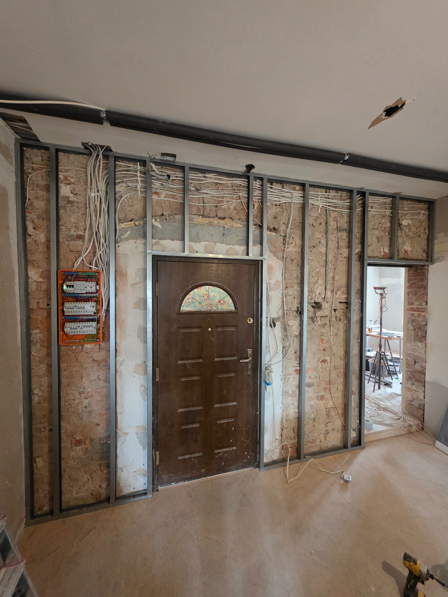 Montaż metalowej konstrukcji ścianki działowej z widoczną instalacją elektryczną i starą cegłą. W tle drzwi i widok na kolejne pomieszczenie w trakcie prac.