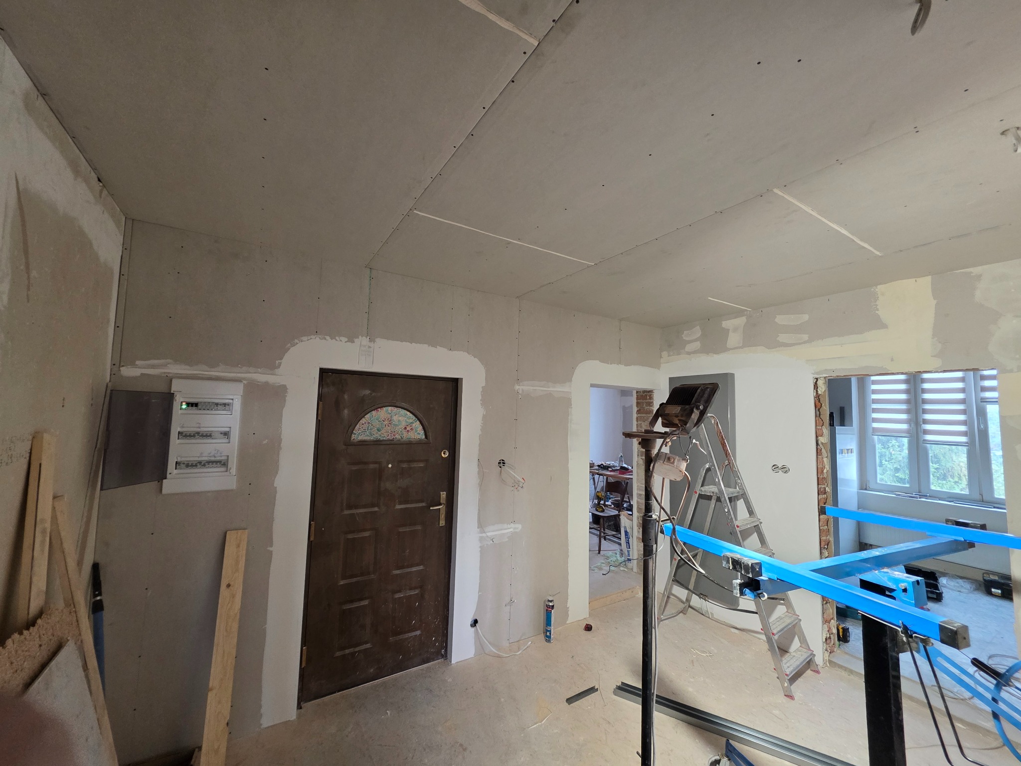 Wnętrze w trakcie montażu płyt karton-gips na ścianach i suficie. Widoczna drabina, lampa budowlana i narzędzia. Częściowo wykończone ściany, surowy stan.