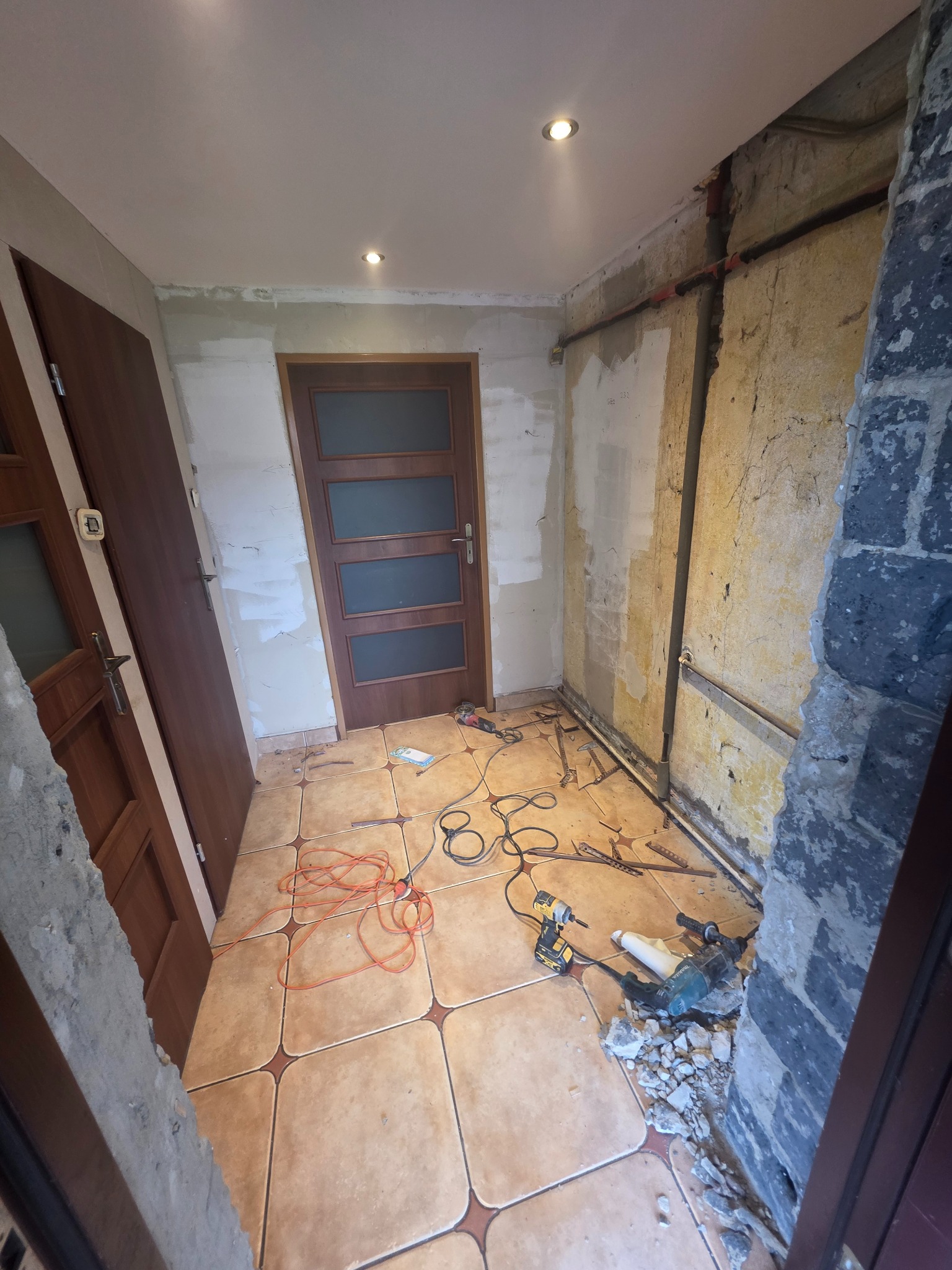 Wnętrze w trakcie demontażu: odsłonięte ściany, narzędzia na podłodze z płytek, widoczne instalacje. Dwa rodzaje drzwi w pomieszczeniu. Prace przygotowawcze do remontu.