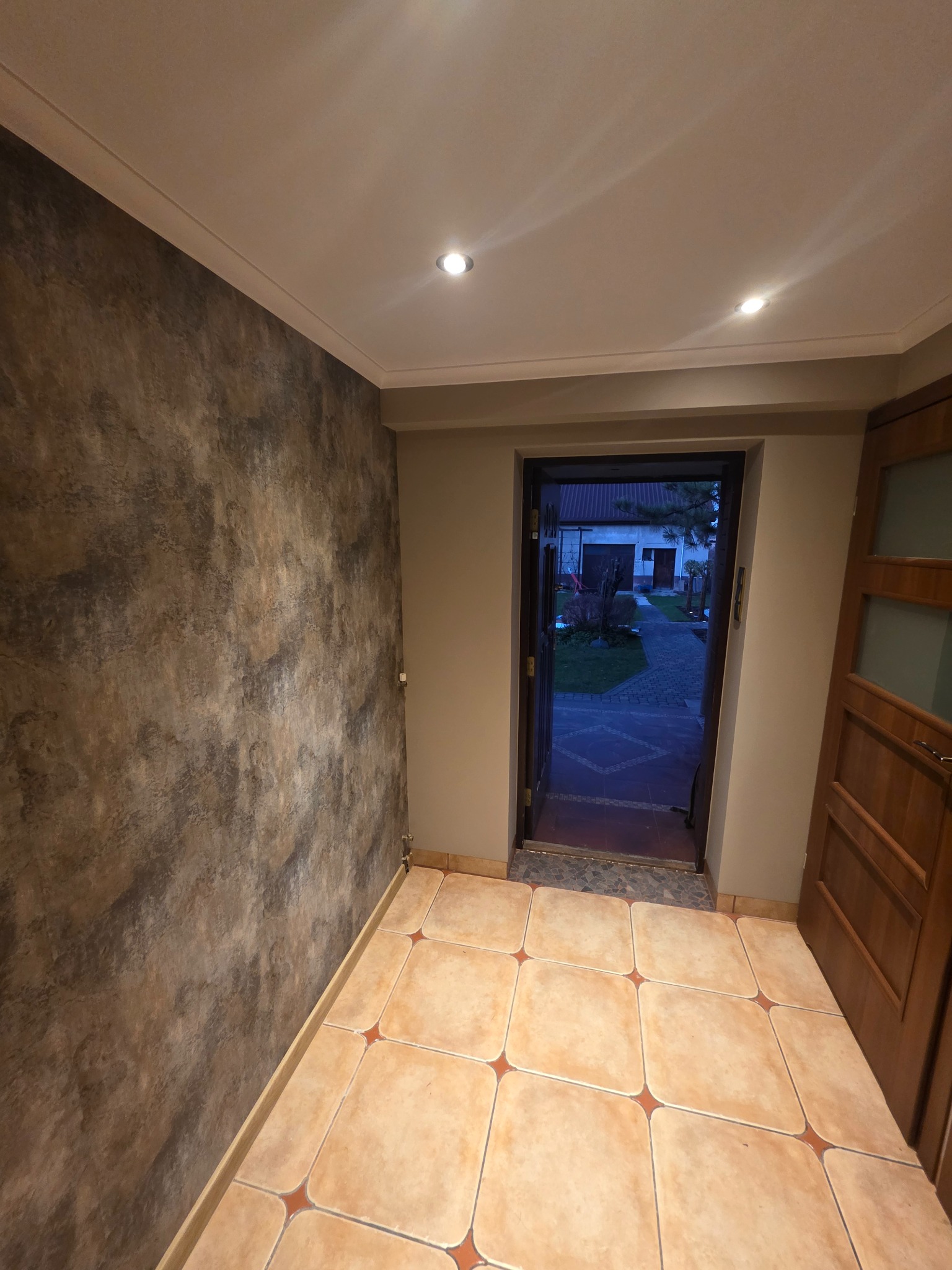 Korytarz po remoncie: dekoracyjna ściana w odcieniach brązu i beżu, beżowa podłoga z kwadratowych płytek z czerwonymi akcentami, drewniane drzwi i widok na ogród.