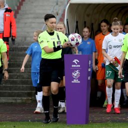 Powerful Mind Patrycja Turczyn - Sędziowie piłkarscy w neonowych koszulkach prowadzą zawodniczki na boisko. Na pierwszym planie fioletowy postument z logo Orlen Ekstraligi.