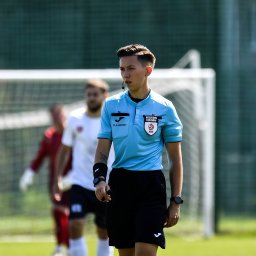 Powerful Mind Patrycja Turczyn - Młody sędzia piłkarski w błękitnej koszulce z odznaką, czarnych spodenkach i sportowym zegarku na boisku podczas meczu. W tle siatka bramki i zawodnicy.