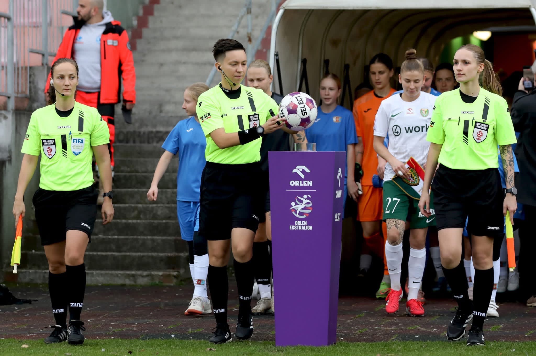 Sędziowie piłkarscy w neonowych koszulkach prowadzą zawodniczki na boisko. Na pierwszym planie fioletowy postument z logo Orlen Ekstraligi.