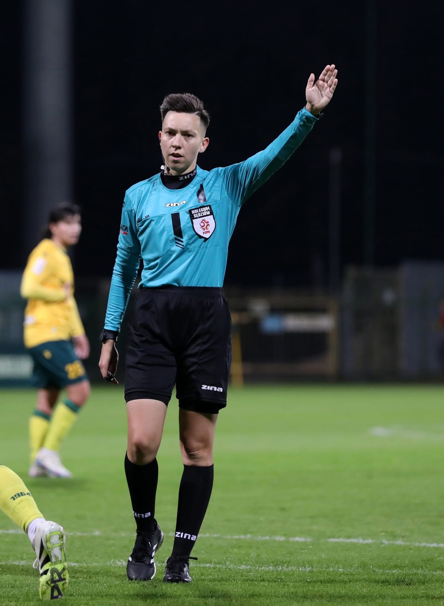 Sędzia piłkarski w turkusowej koszuli i czarnych spodenkach sygnalizuje przerwę w grze, unosząc rękę na tle zielonej murawy i zawodników w oddali.