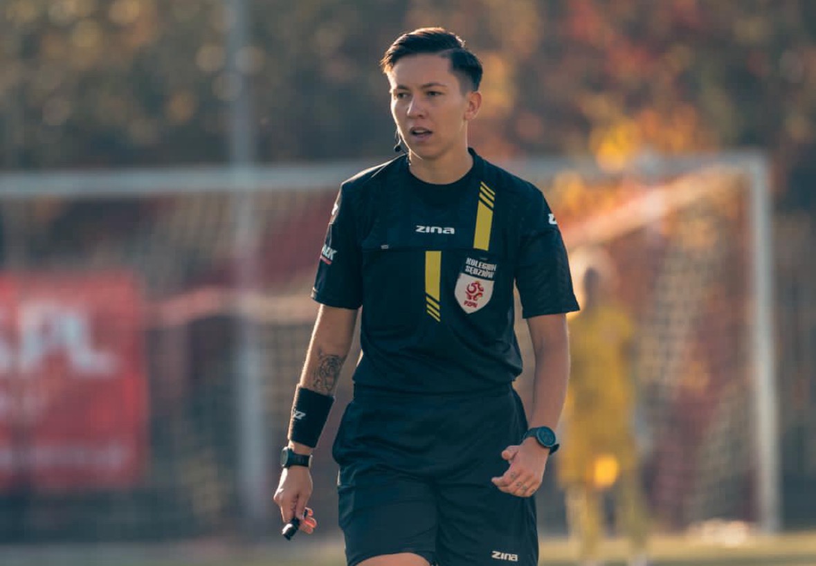 Kobieta w stroju sędziego piłkarskiego na tle boiska, z widocznym gwizdkiem w dłoni i tatuażem na ramieniu, w trakcie meczu.