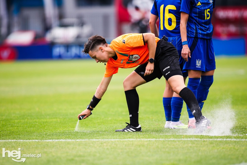 Arbiter w pomarańczowej koszulce, pochylony, zaznacza linię na zielonej murawie boiska, obok zawodniczki w niebieskich strojach. Widoczny zegarek sportowy.