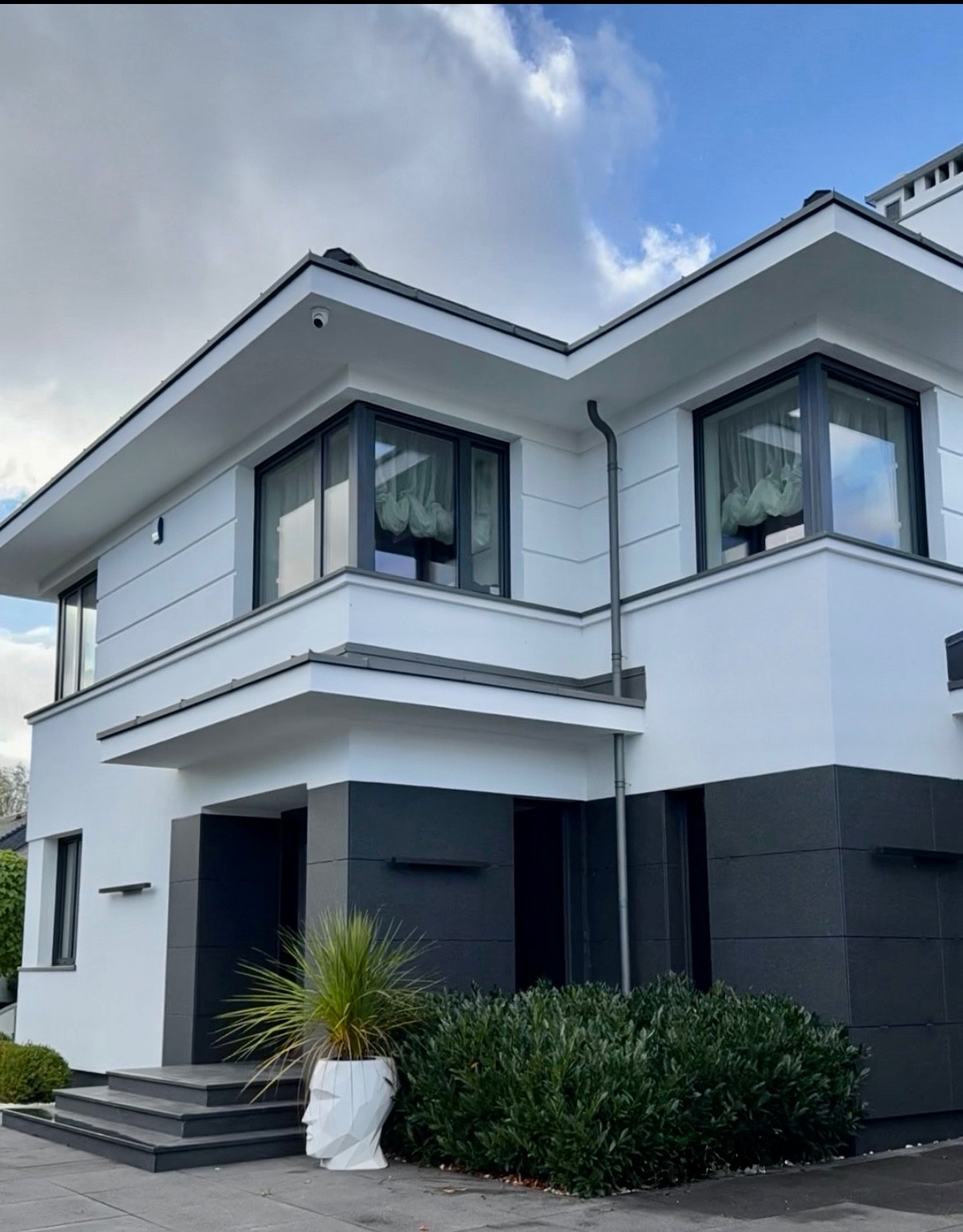 Nowoczesny dom z biało-grafitową elewacją, minimalistycznym designem i dużymi oknami. Uwagę przykuwa donica w kształcie głowy i starannie utrzymana zieleń przed wejściem.