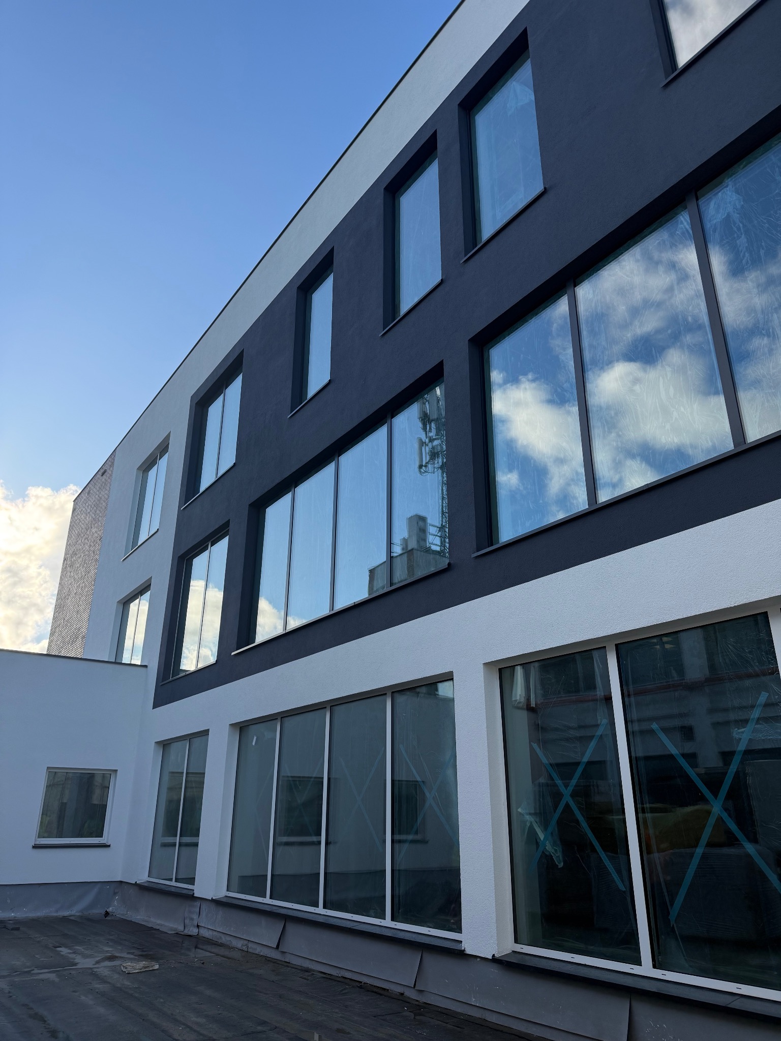 Dwukolorowa elewacja budynku z dużymi oknami, w których odbija się niebo. Widoczne zabezpieczenia okien folią i taśmą. Ujęcie z dołu, błękitne niebo w tle.