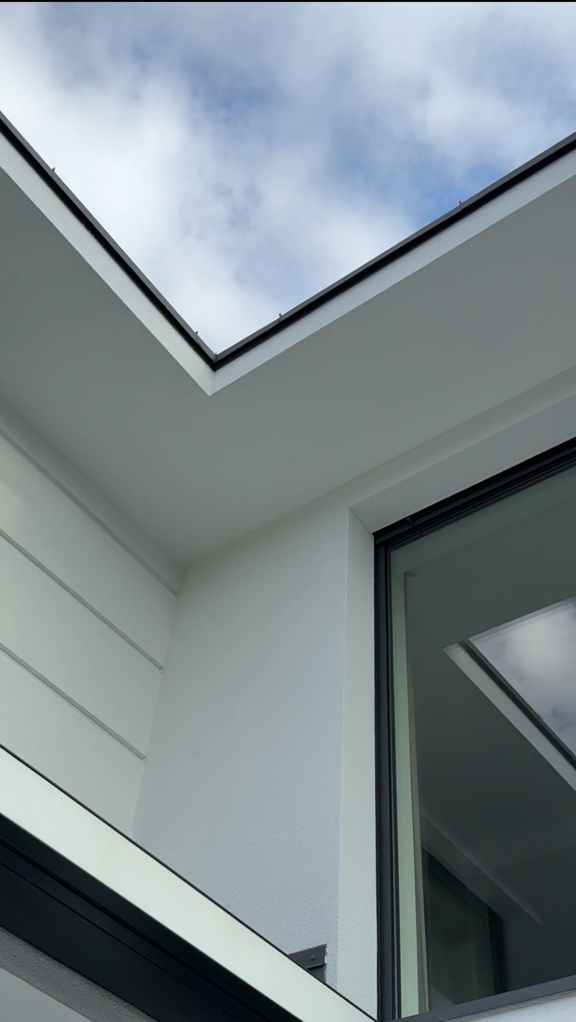 Nowoczesna elewacja domu z białym tynkiem strukturalnym, czarnymi detalami okien i dachu. Widok z dołu na fragment ściany i niebo.