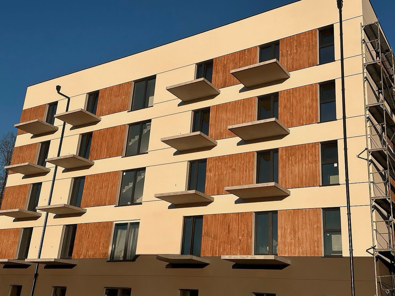 Nowoczesny budynek wielorodzinny z beżową elewacją, drewnianymi panelami i balkonami. Widoczne rusztowania z boku budynku. Słoneczny dzień, błękitne niebo.