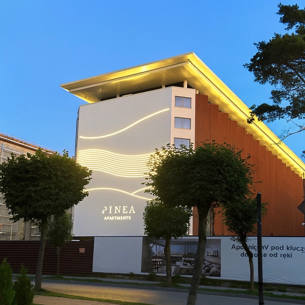 Nowoczesny budynek apartamentowy Pinea z oświetloną elewacją, drewnianymi panelami i złotym dachem, widziany z poziomu ulicy o zmierzchu.