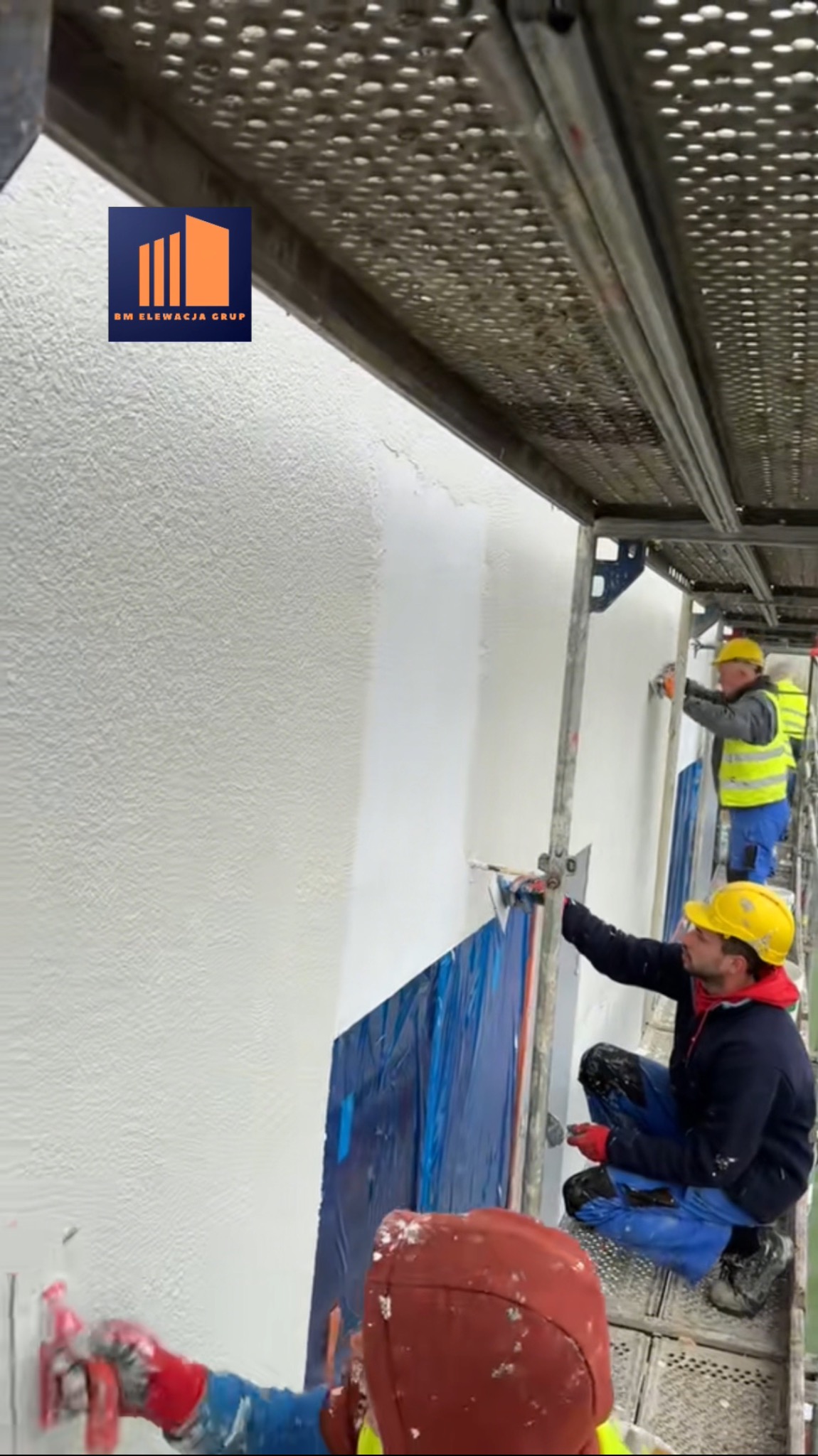 Praca na rusztowaniu: ekipa tynkuje elewację budynku na biało. Część ściany zabezpieczona folią. Widoczny fragment logo firmy na górze kadru.