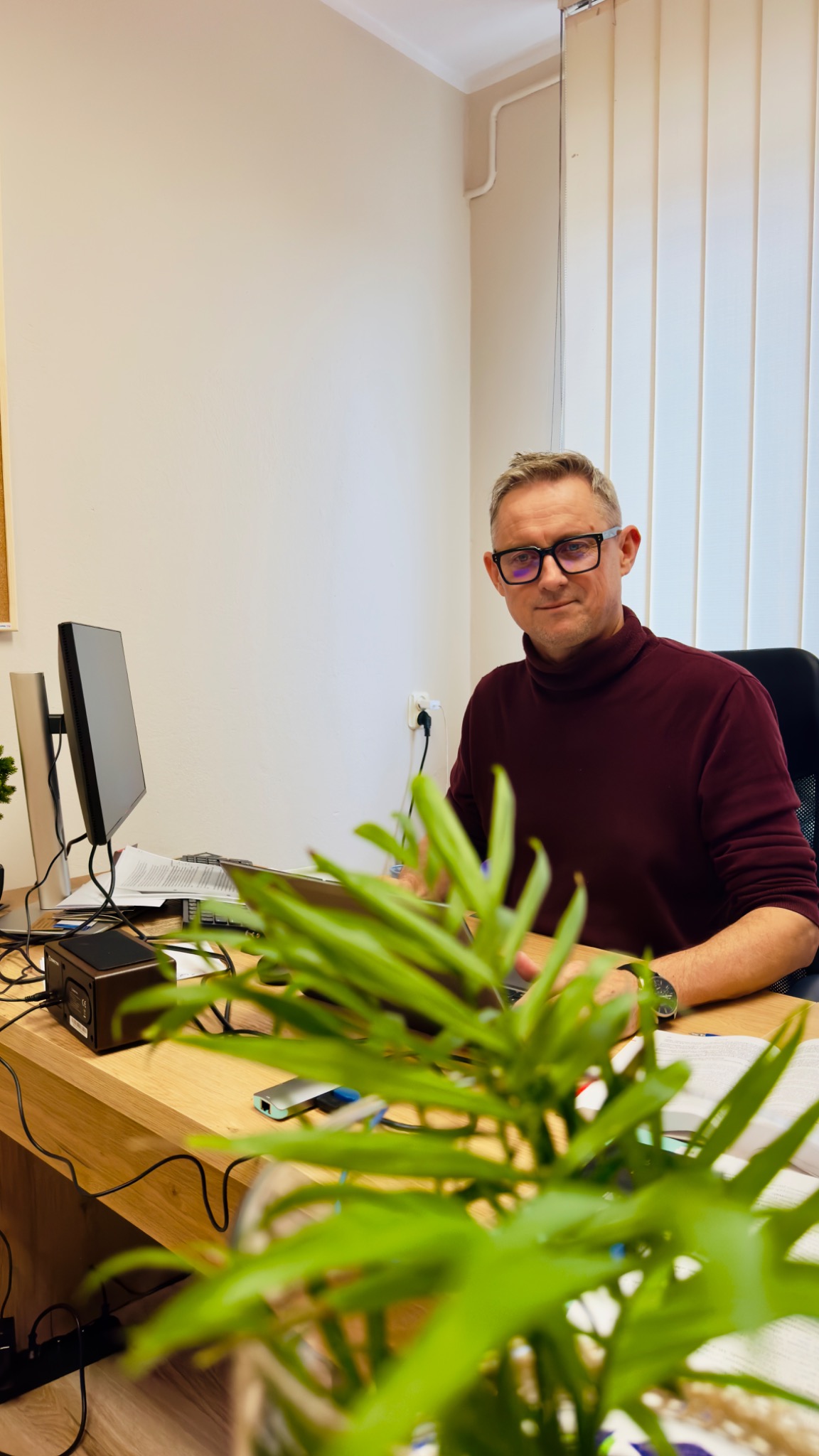 Mężczyzna w okularach siedzi za biurkiem z komputerem i dokumentami, z rośliną doniczkową na pierwszym planie. Neutralne tło biurowe z żaluzjami.