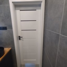 Firma Remontowo- Budowlana Easy Spray Tomasz Dziubanowski - Nowoczesna, biała, wewnętrzna stolarka drzwiowa z czarnymi akcentami w łazience wykończonej szarą płytką. Widoczny fragment umywalki z szafką.