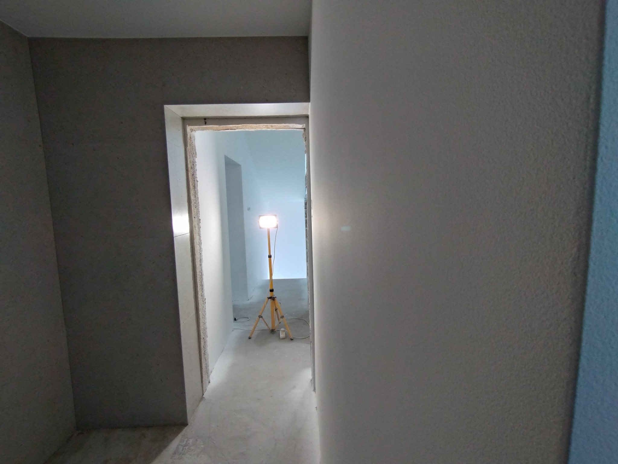 Wnętrze w trakcie malowania: szare ściany z widoczną fakturą, przejście do jasnego pomieszczenia oświetlonego lampą na statywie. Prace wykończeniowe w toku.