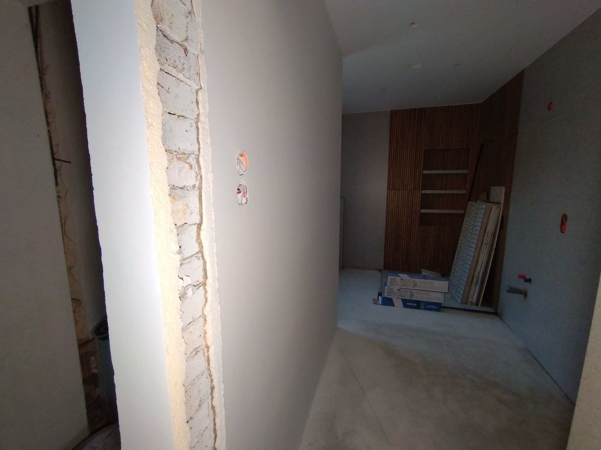 Wnętrze w trakcie malowania: świeżo pomalowana ściana z widocznymi puszkami elektrycznymi, surowa ściana z cegły i przygotowane panele dekoracyjne w tle.