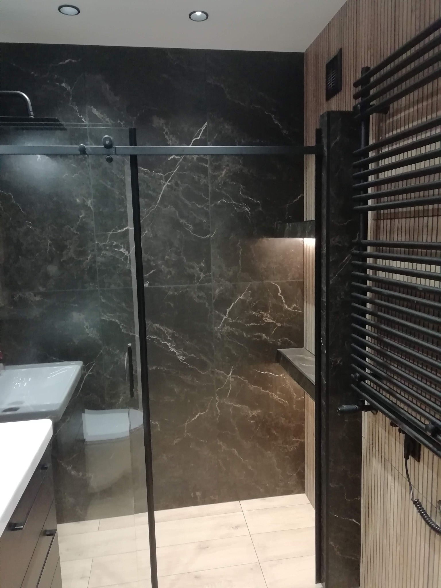 Nowoczesna łazienka z czarnymi płytkami imitującymi marmur, kabiną prysznicową typu walk-in z czarnymi ramami i grzejnikiem drabinkowym w kolorze czarnym.