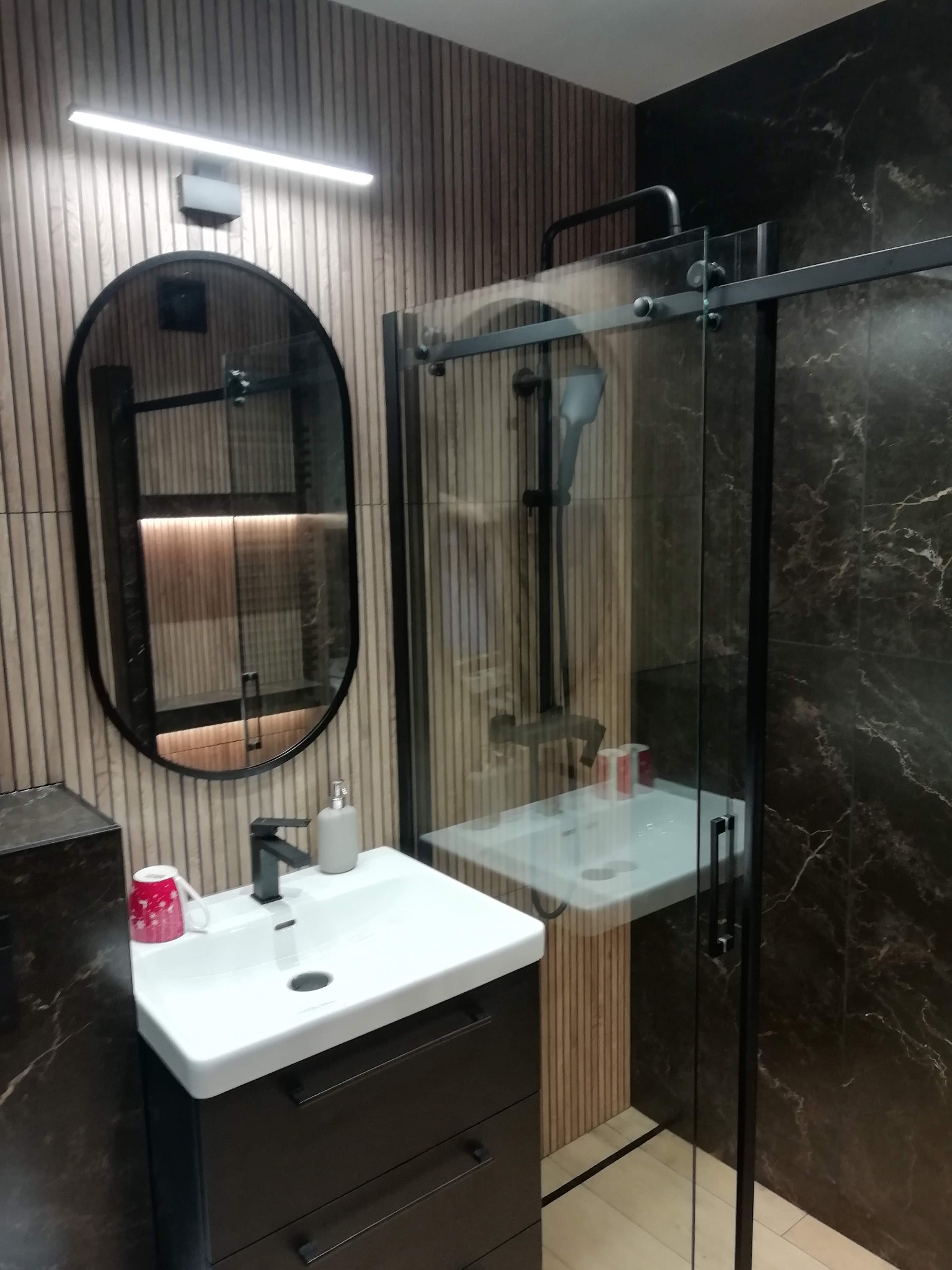 Nowoczesna łazienka z kabiną prysznicową typu walk-in, umywalką nablatową i owalnym lustrem w czarnej ramie. Ściany wykończone panelami i płytkami imitującymi marmur.