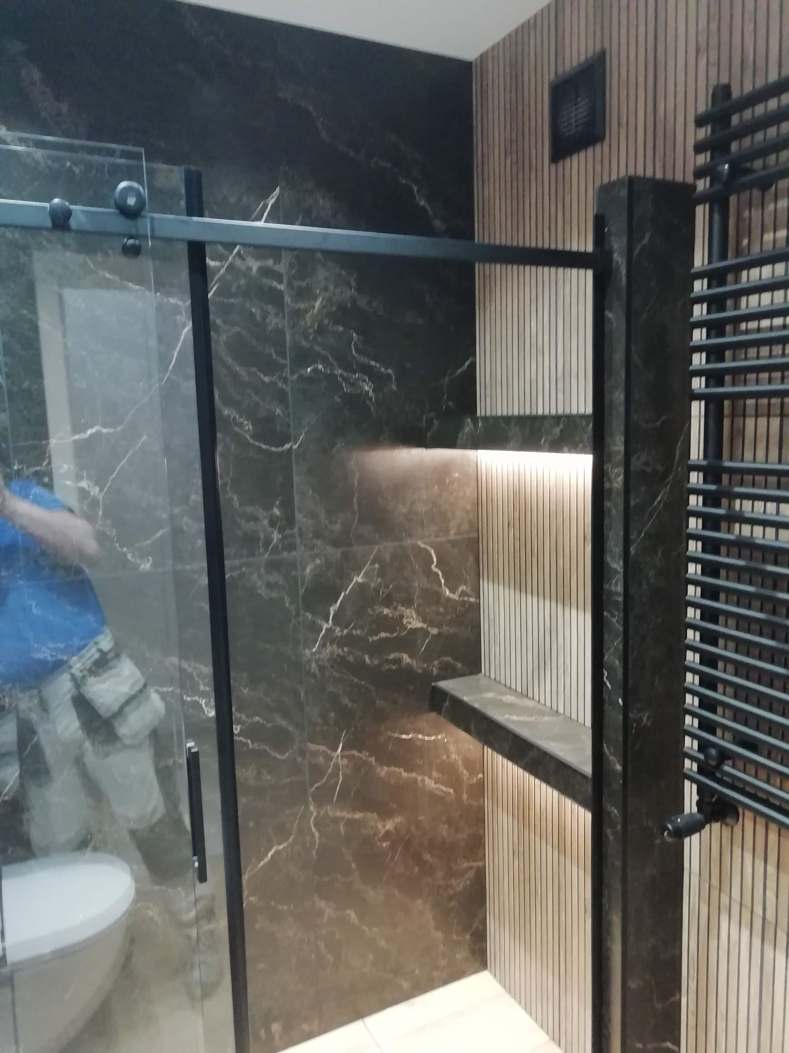 Nowoczesna łazienka z kabiną prysznicową typu walk-in, płytkami imitującymi marmur i drewnianymi lamelami z podświetleniem LED. Grzejnik drabinkowy w kolorze czarnym.