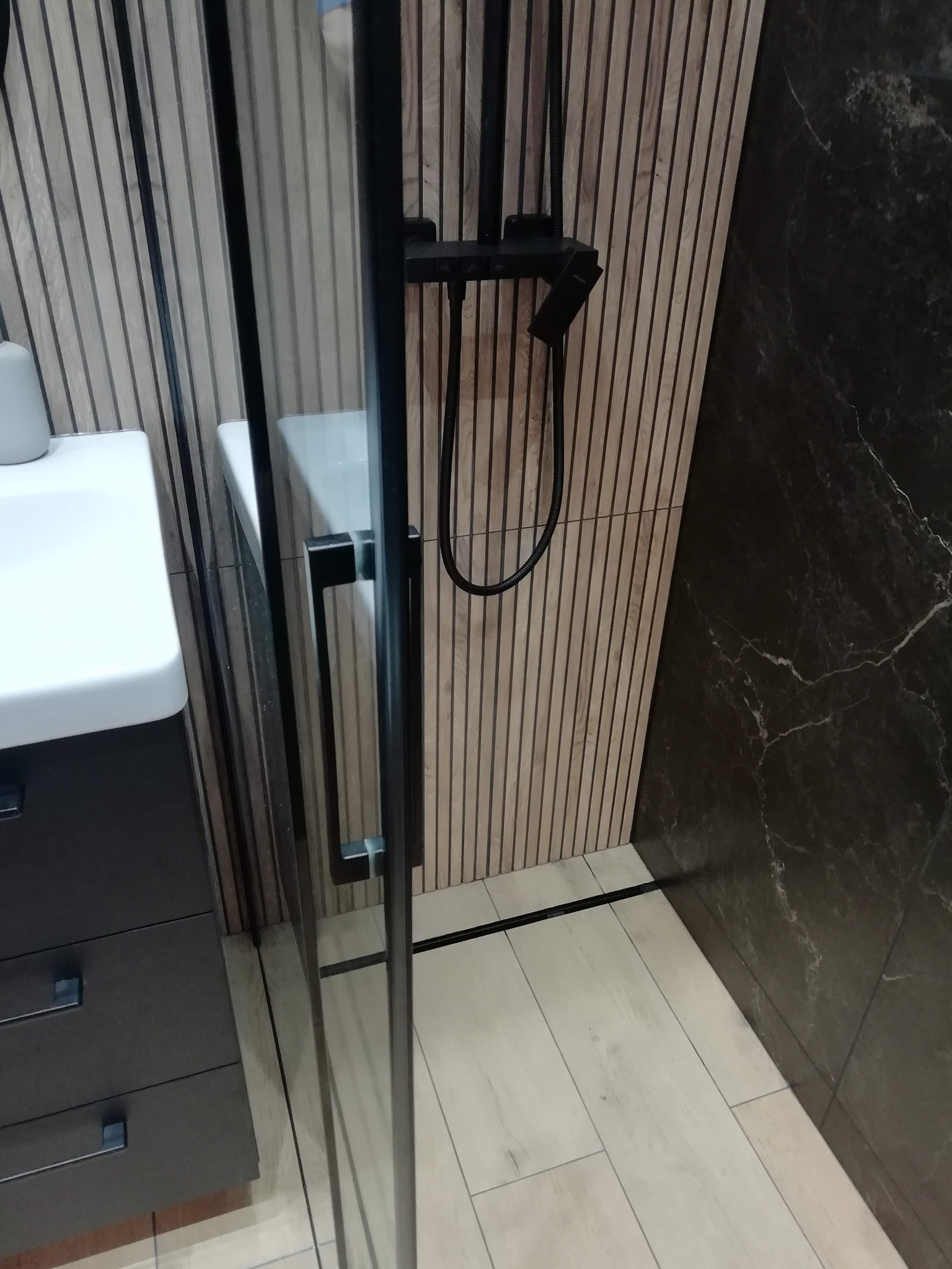 Nowoczesna łazienka z kabiną prysznicową typu walk-in, drewnianymi lamelami na ścianie i czarnymi akcentami armatury. Widoczna umywalka z szafką i marmurowe płytki.