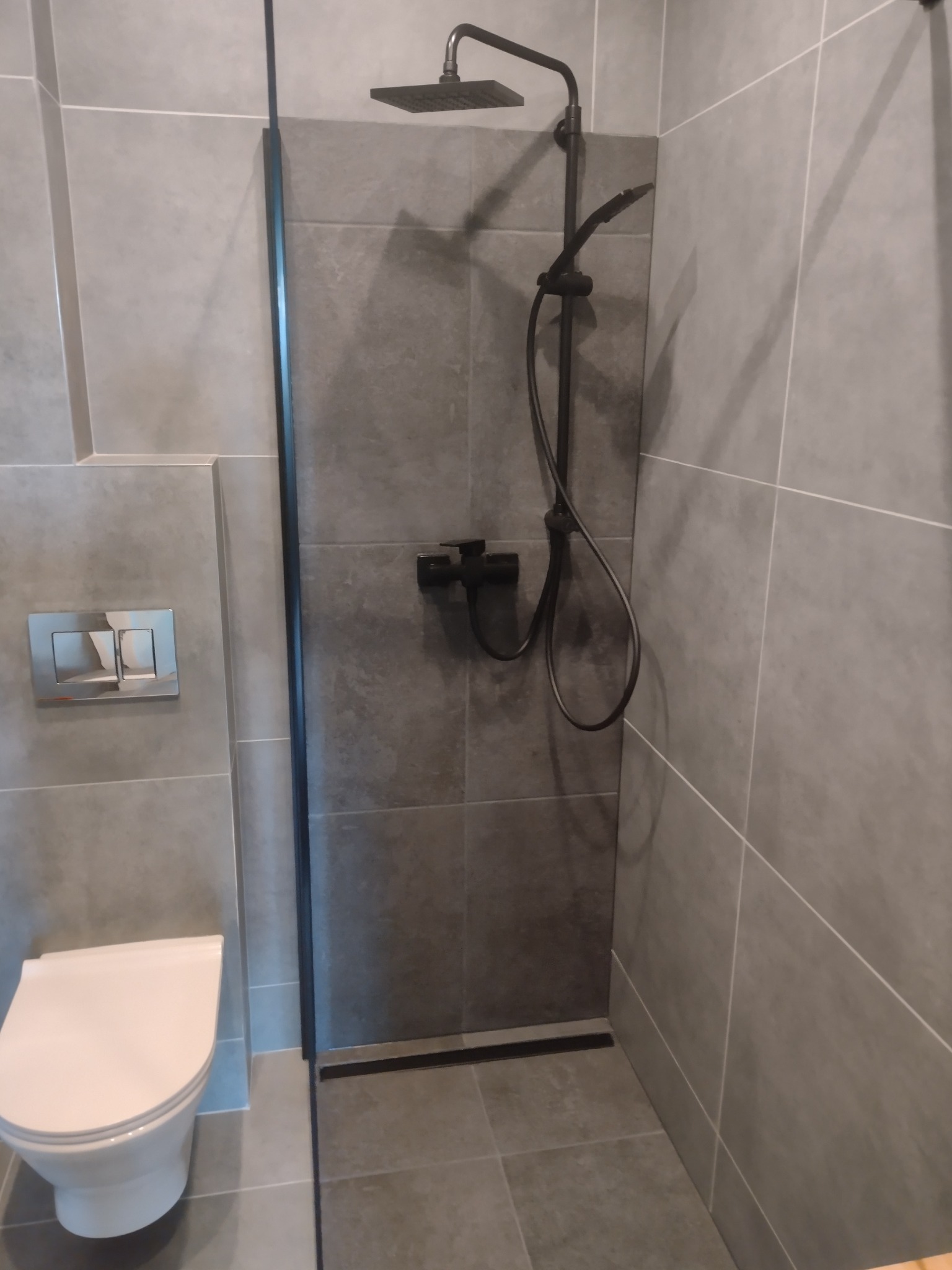 Nowoczesna łazienka z szarymi płytkami, czarną armaturą prysznicową i podtynkową toaletą. Minimalistyczny design z akcentem na funkcjonalność i stonowaną kolorystykę.