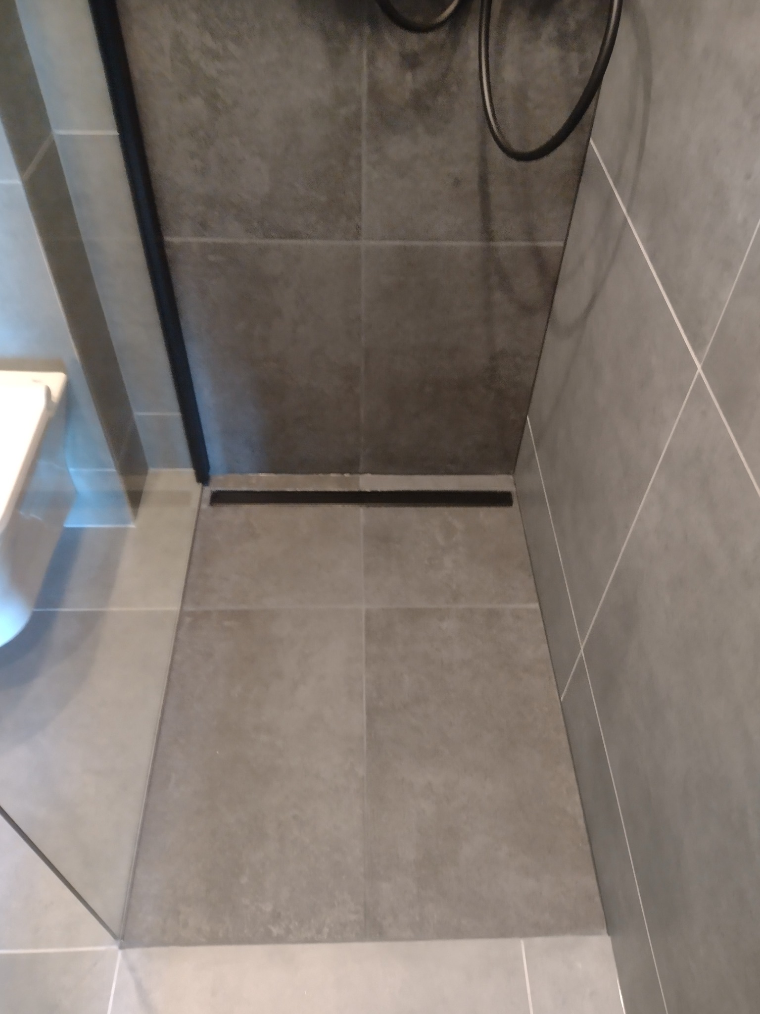 Nowoczesna łazienka z szarymi płytkami, widoczny odpływ liniowy i fragment kabiny prysznicowej. Minimalistyczny design, stonowana kolorystyka.