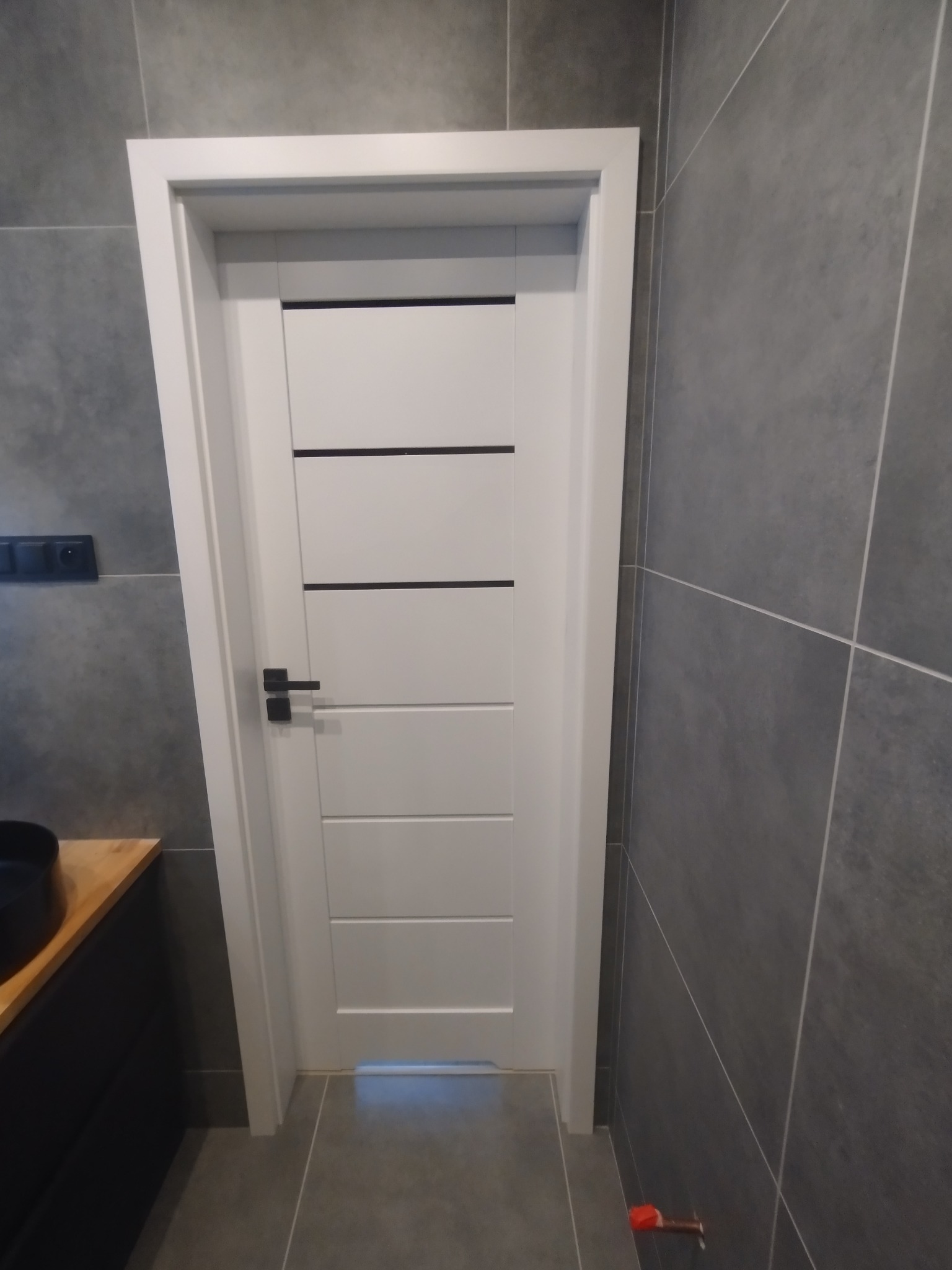 Nowoczesna, biała, wewnętrzna stolarka drzwiowa z czarnymi akcentami w łazience wykończonej szarą płytką. Widoczny fragment umywalki z szafką.