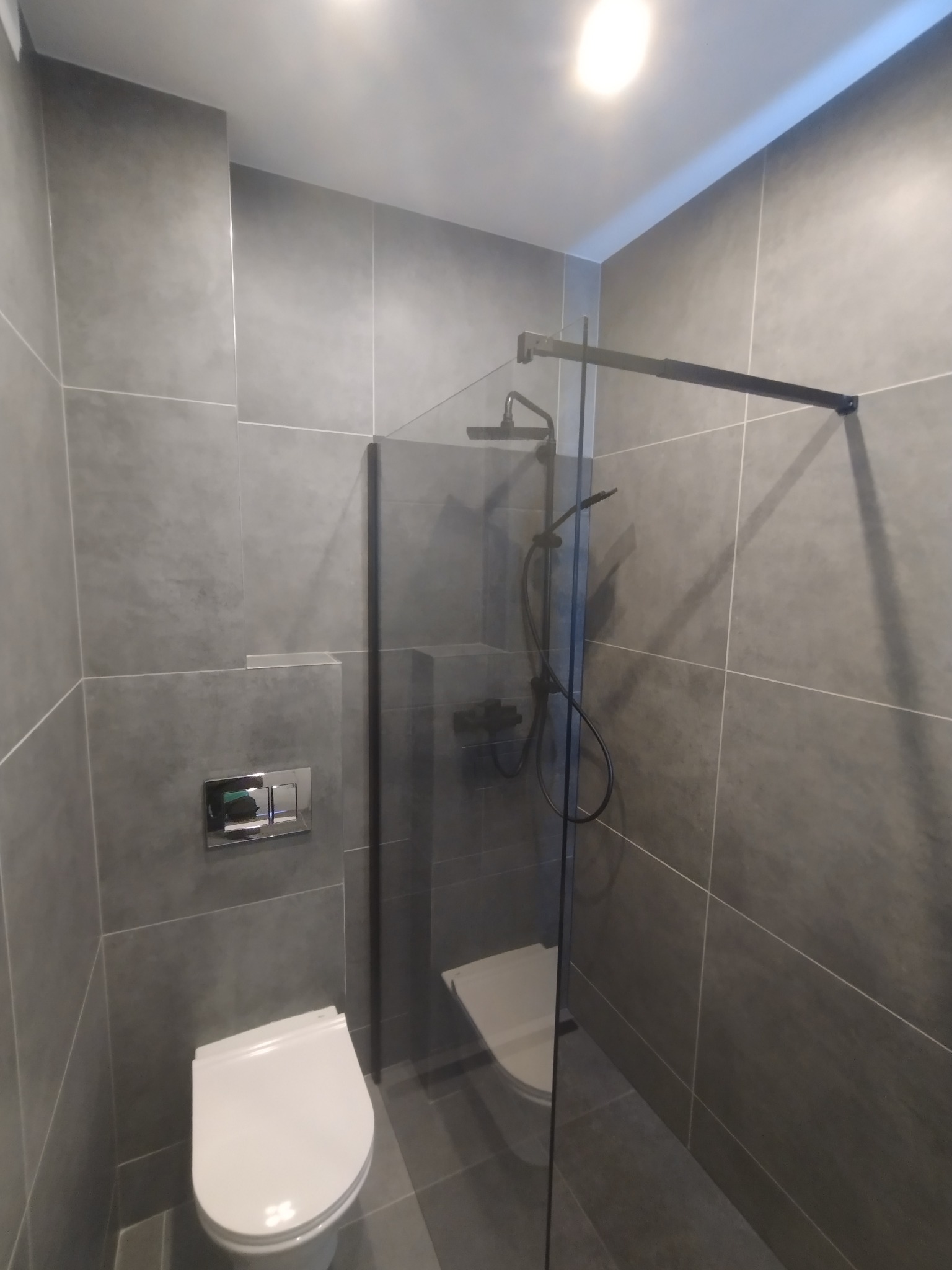 Nowoczesna łazienka w szarościach z kabiną typu walk-in i podtynkową armaturą. Minimalistyczny design, płytki imitujące beton, oświetlenie punktowe.